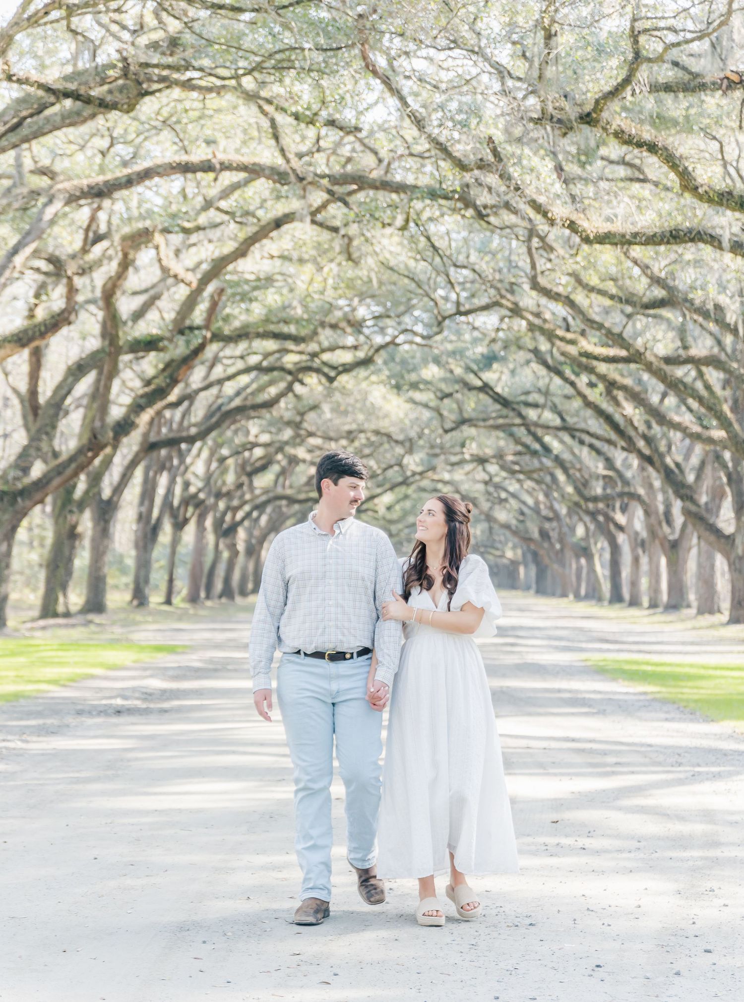 A couple shares a joyful moment while walking down a path lined with majestic oak trees.