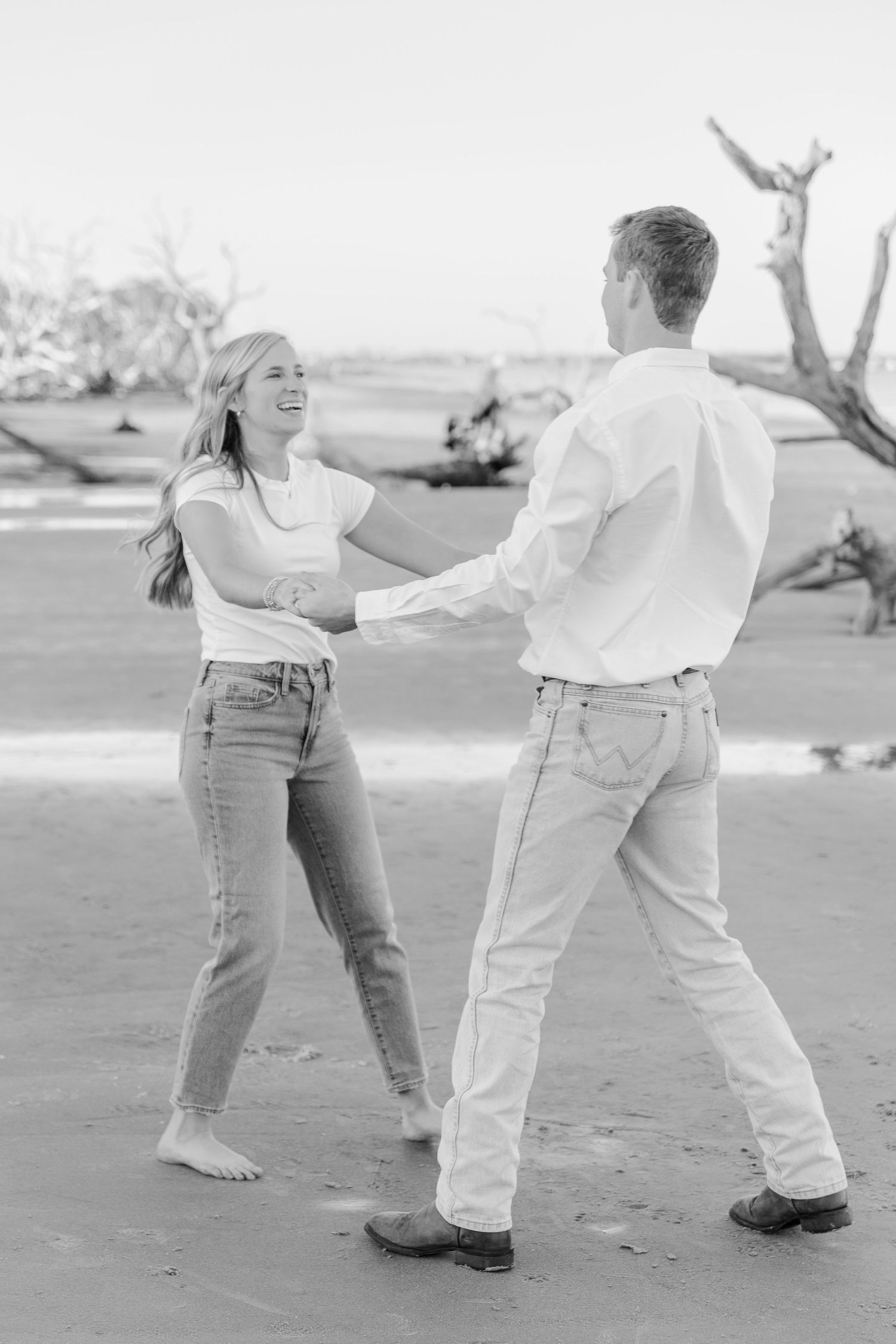 Couple dancing together on a beach with driftwood in the background during a casual photoshoot.