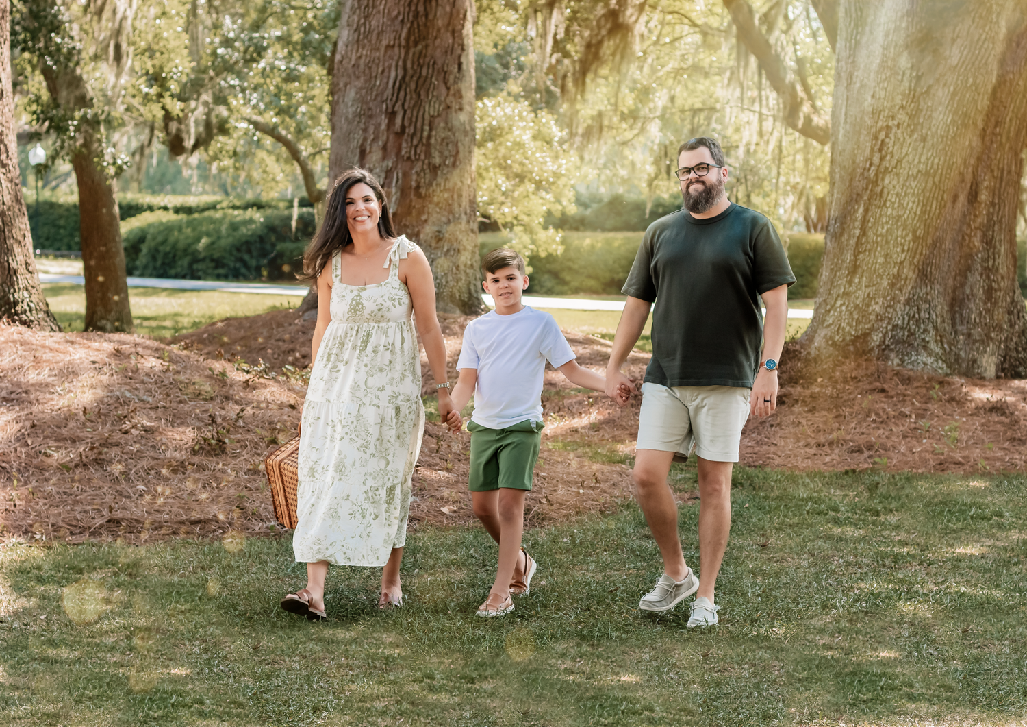 A family walks together on green grass beneath towering oak trees in a sunlit park setting.