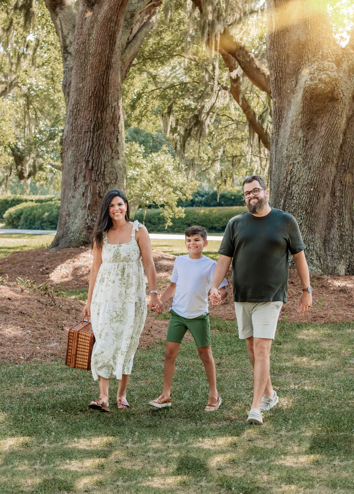 Family of three walks together on grass under large oak trees with picnic basket.