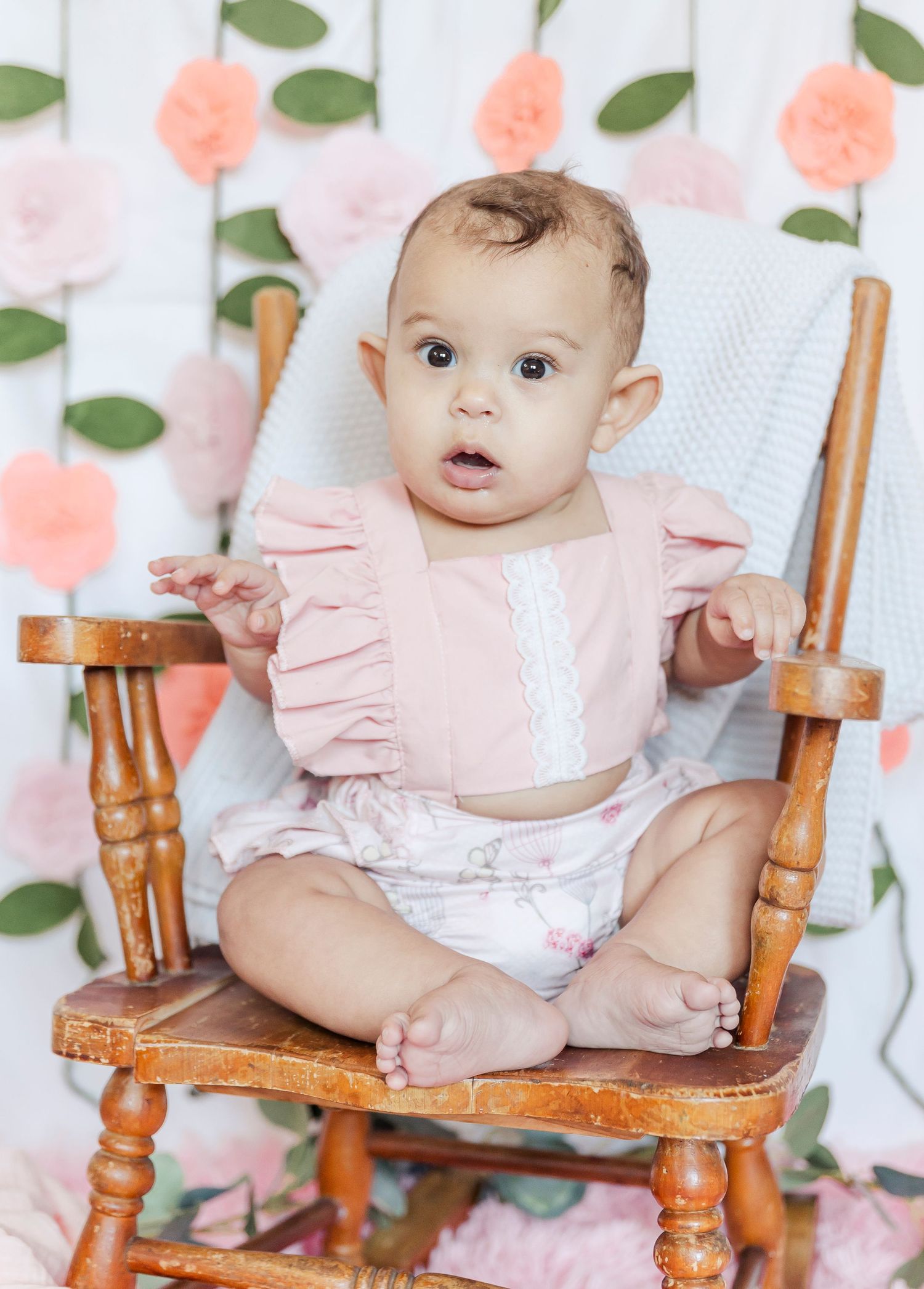 Baby sits in wooden chair wearing pink ruffled outfit against floral wall backdrop.