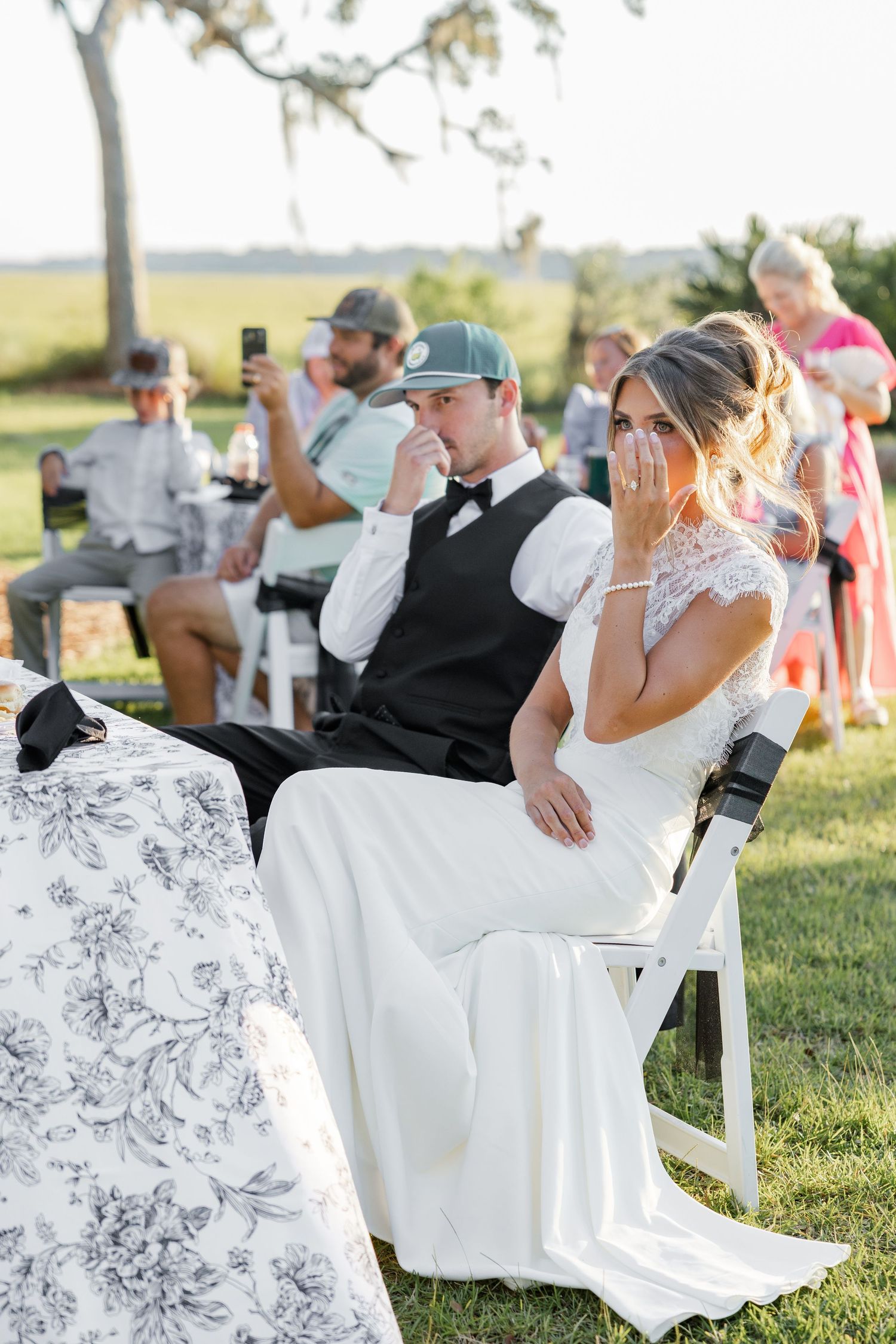 Emotional bride and groom wipe tears during wedding speeches at outdoor garden reception.