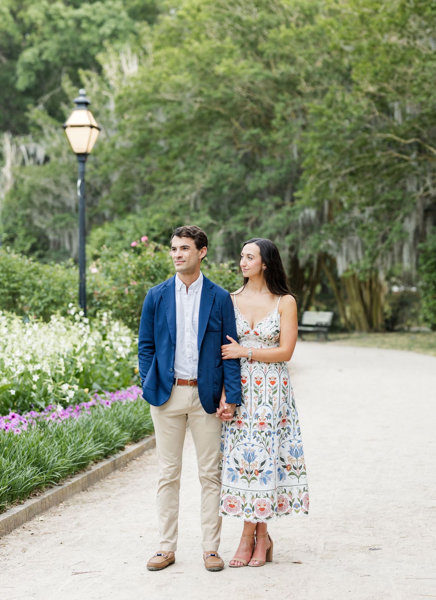 Engaged couple holding hands on a garden path, woman in floral dress and man in navy blazer, lush greenery behind them.