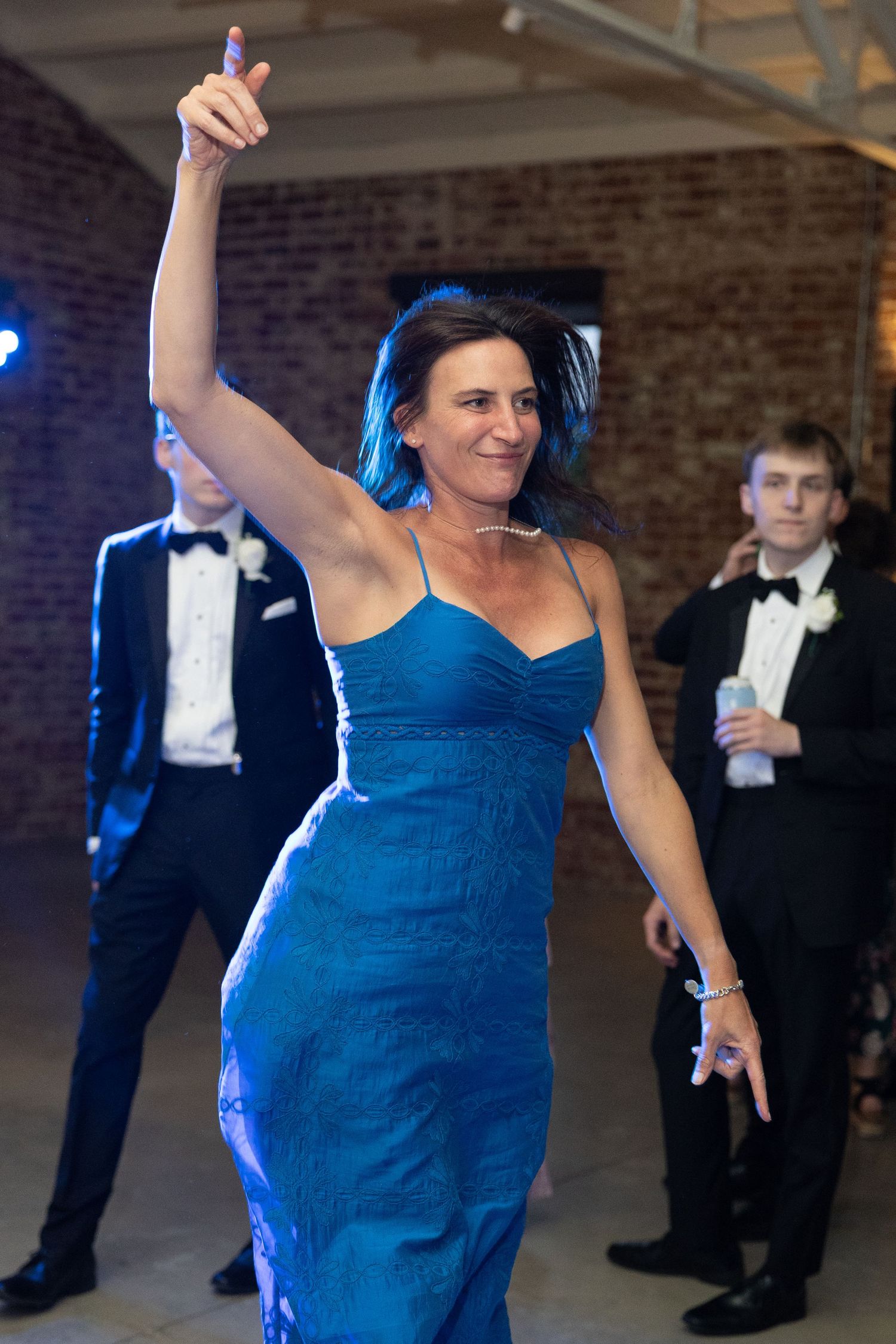 Woman in blue lace gown dances energetically with arm raised at elegant wedding reception.