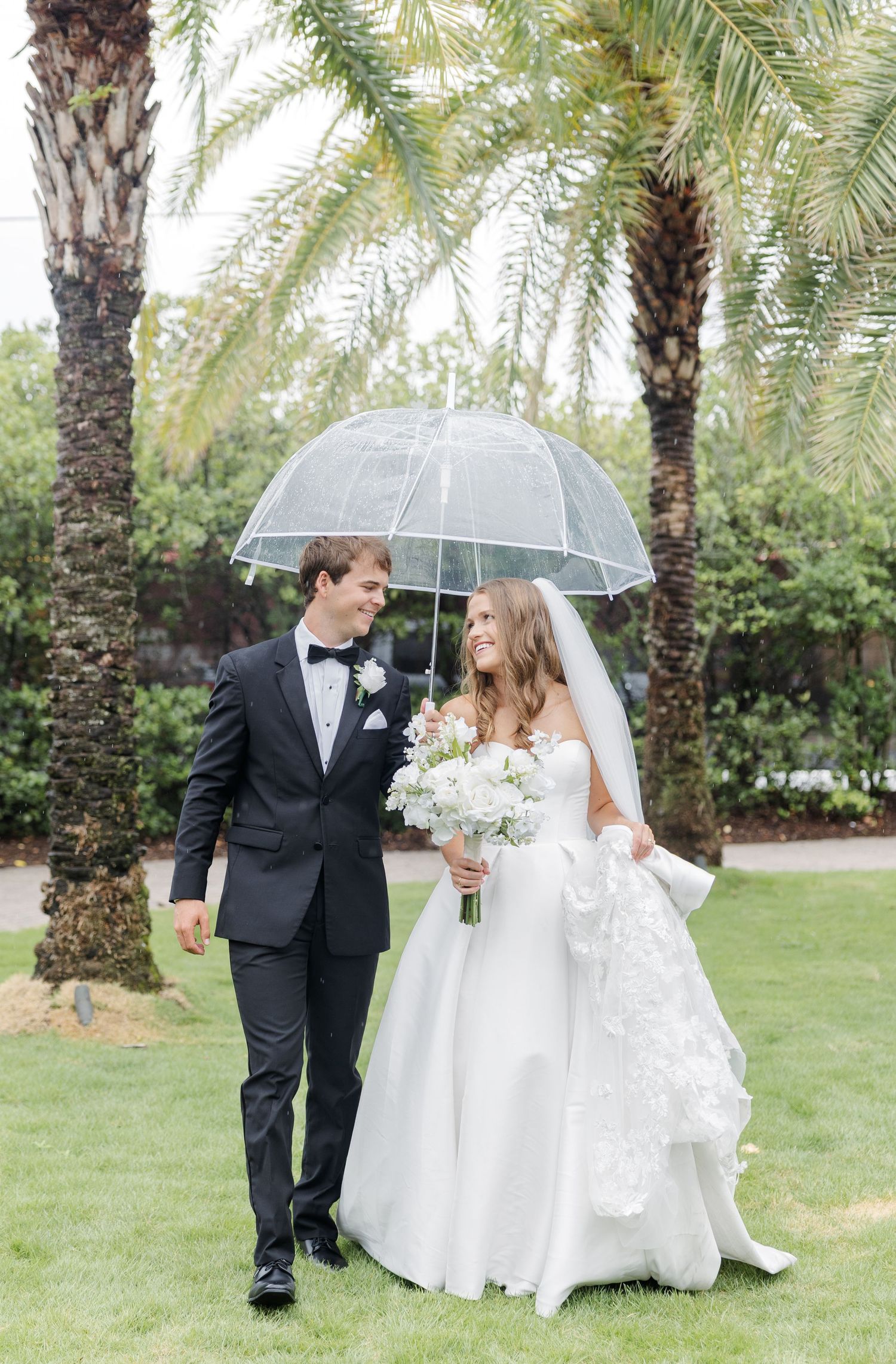 Bride and groom walk under clear umbrella on rainy wedding day with palm trees and white floral bouquet.