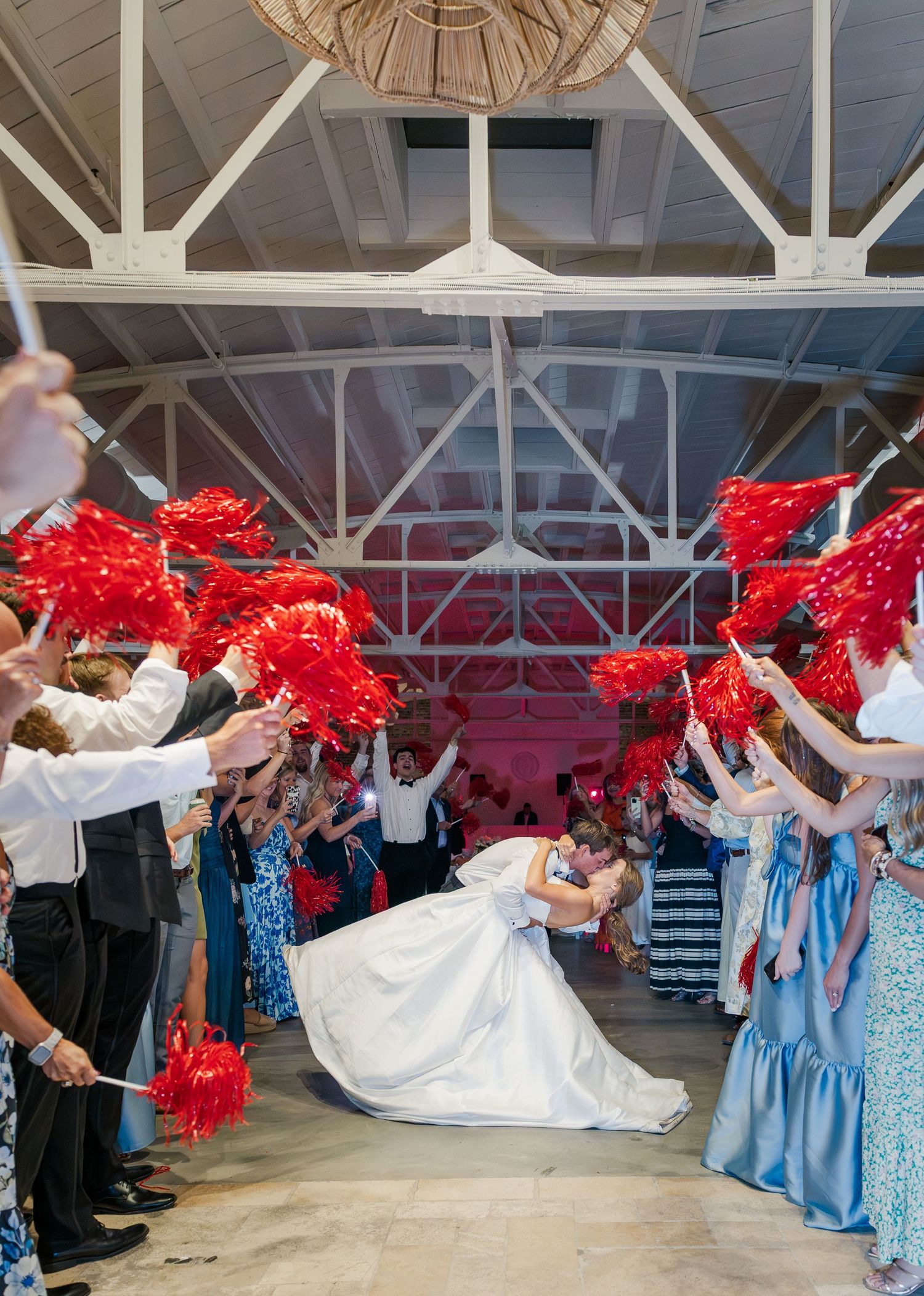 Bride and groom share a dip kiss exit through a tunnel of guests waving red pom-poms at reception.