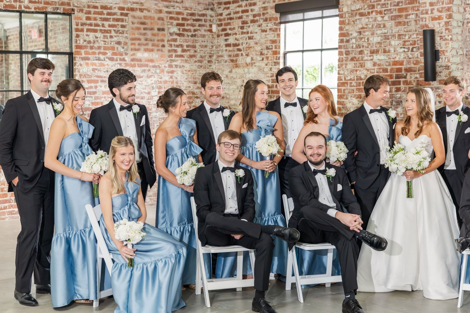 Wedding party in blue bridesmaid dresses and black tuxedos posing playfully in a brick venue.