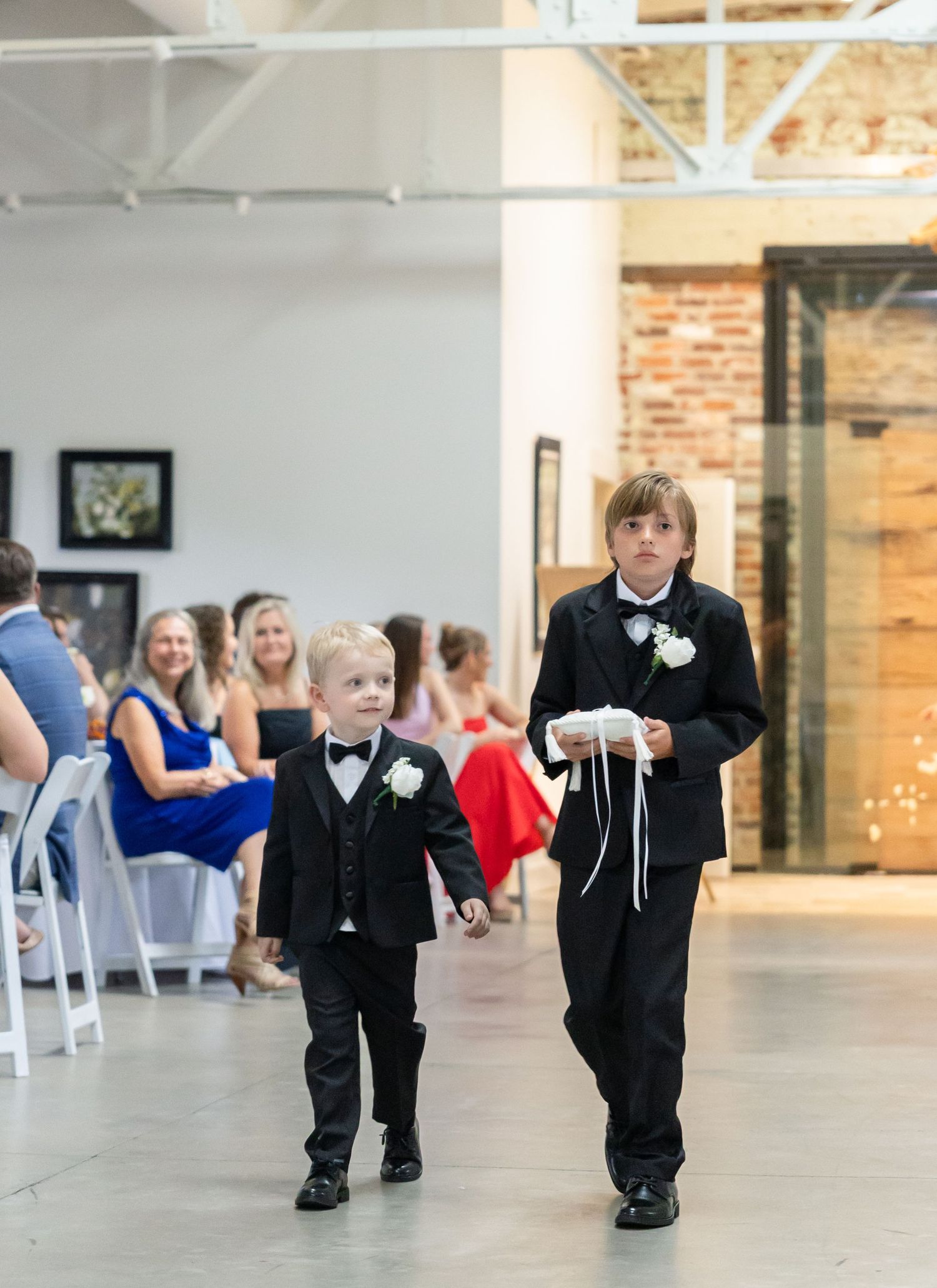 Two young ring bearers in black tuxedos walk down the aisle during a wedding ceremony.