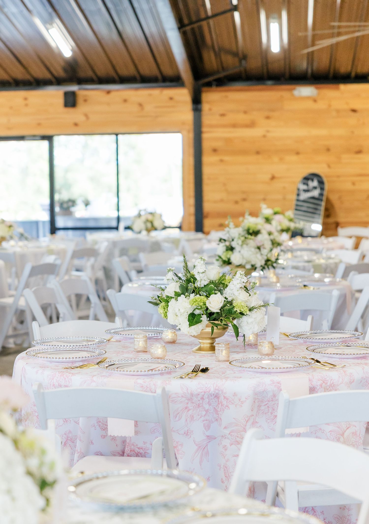 Elegant wedding reception hall with round tables, pink toile linens, white floral centerpieces, and gold accents.