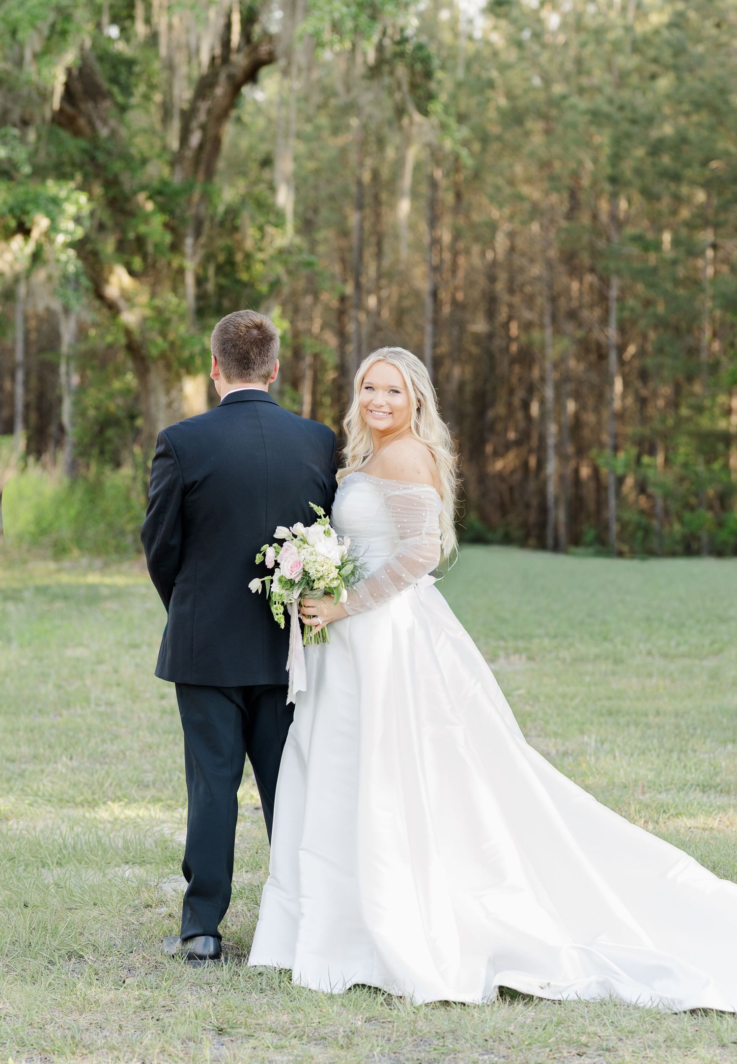 Bride in off-shoulder ball gown holding pink bouquet looks back at camera with groom outdoors.