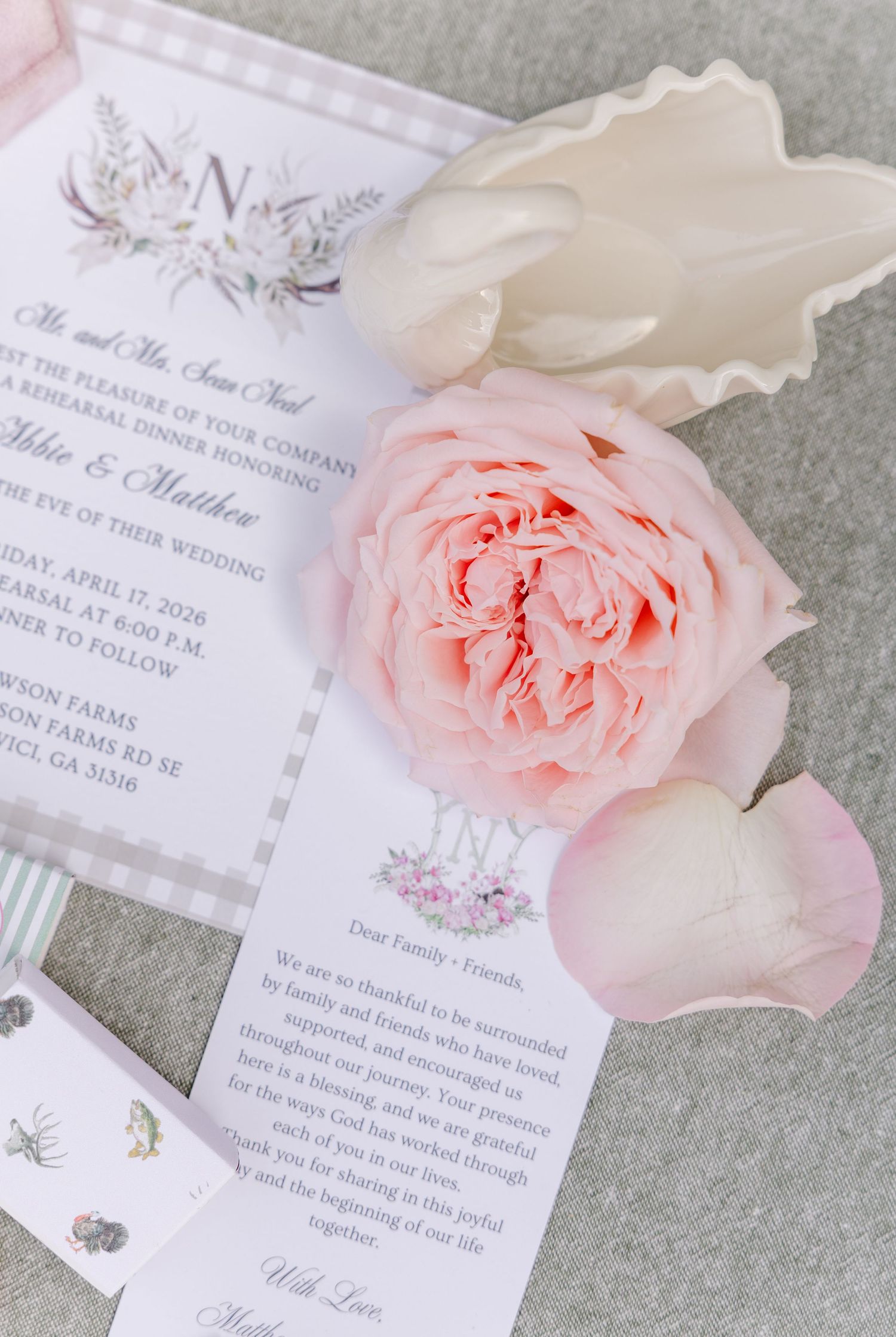 Pink garden rose and white flower rest on elegant rehearsal dinner invitation with floral monogram design.