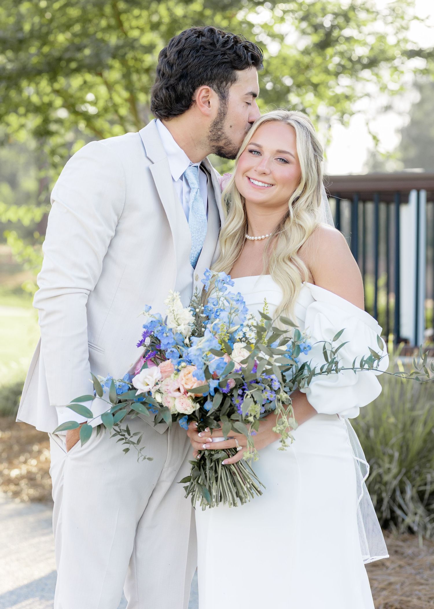 Bride holding blue and white bouquet smiles as groom in tan suit kisses her cheek outdoors.
