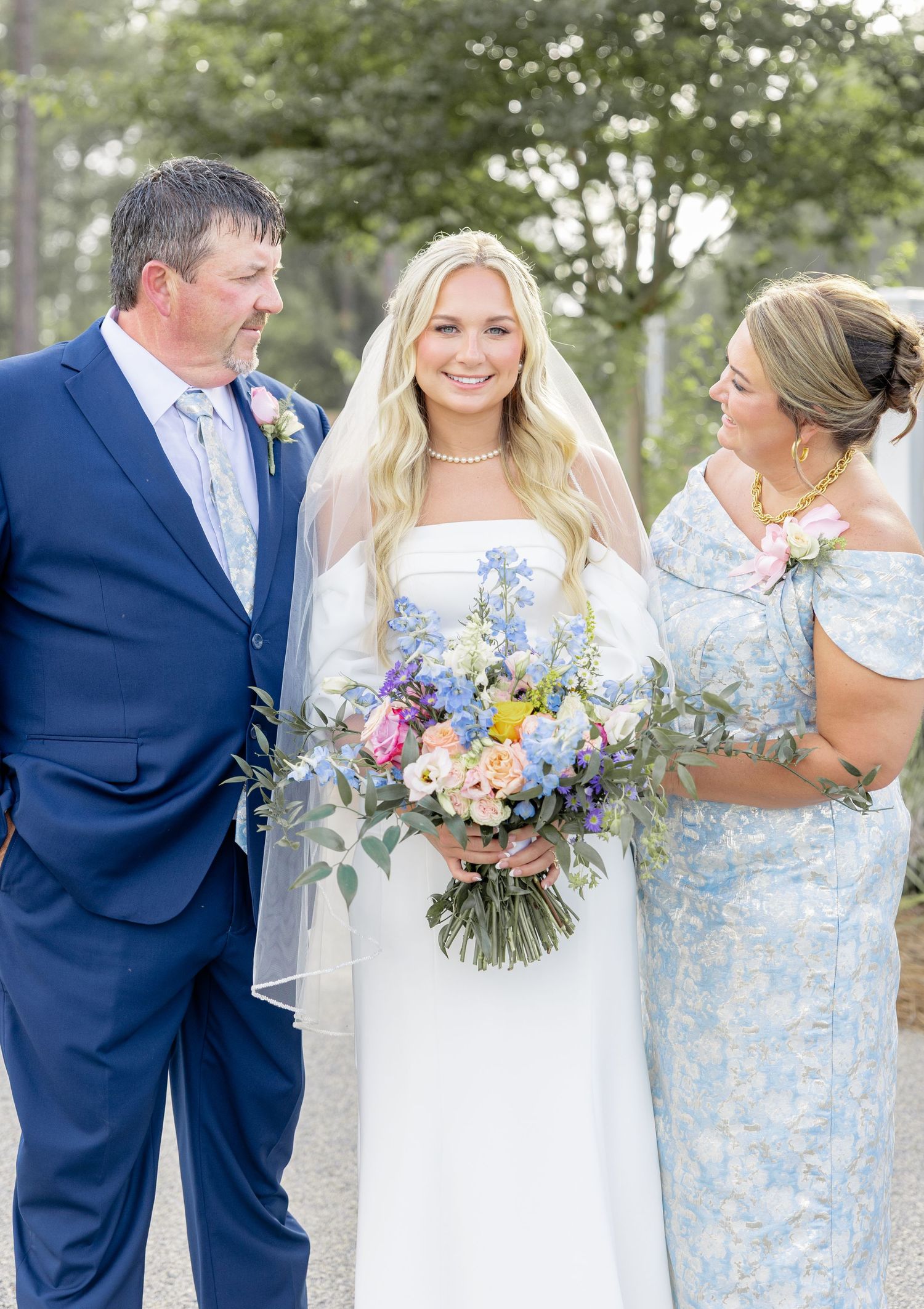 Bride in white gown holding colorful bouquet poses with parents on her wedding day.