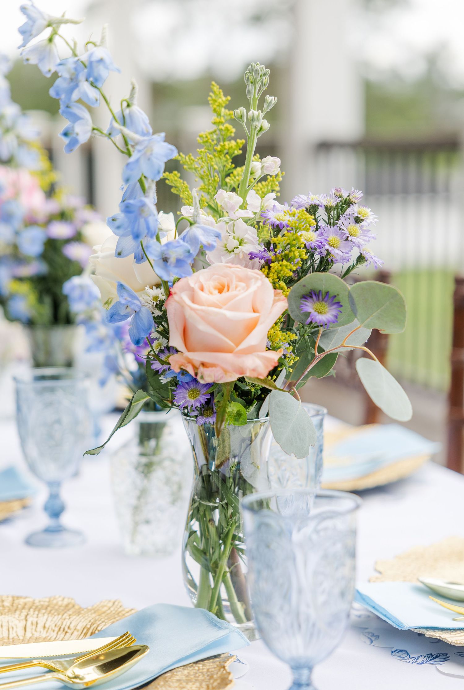 Elegant wedding table centerpiece with peach rose, blue delphinium, and purple wildflowers in a glass vase.