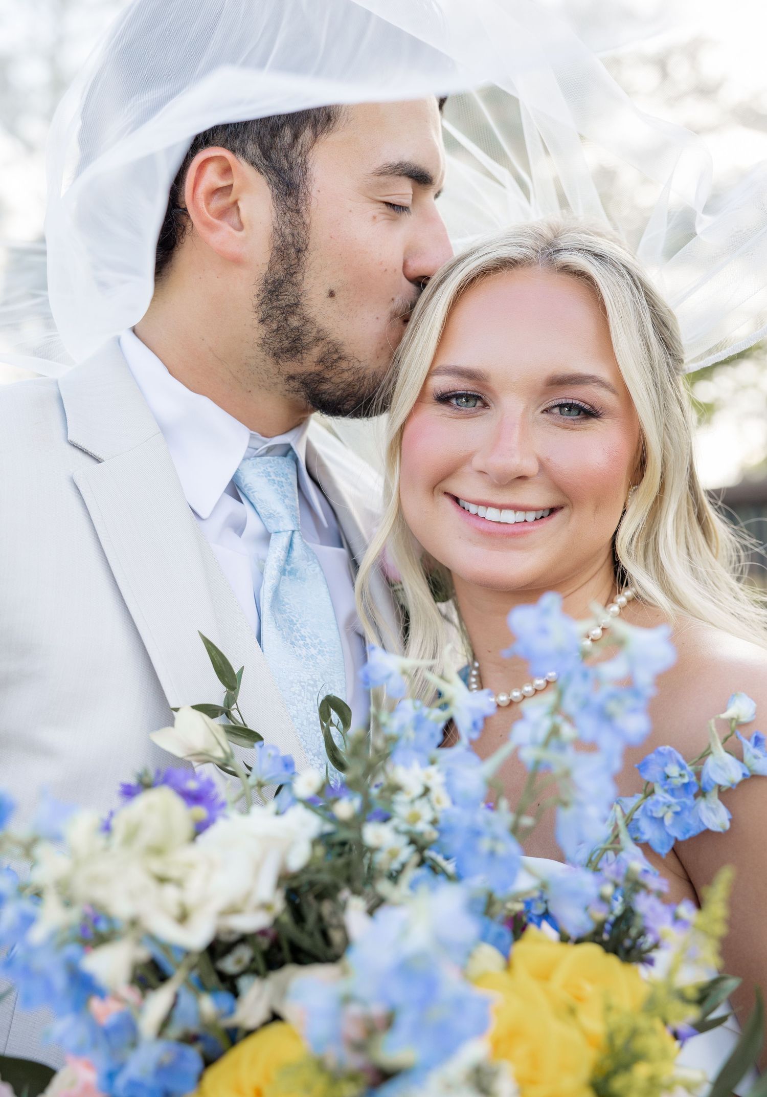 Groom kisses bride on cheek as she smiles holding blue and yellow wedding bouquet under veil.
