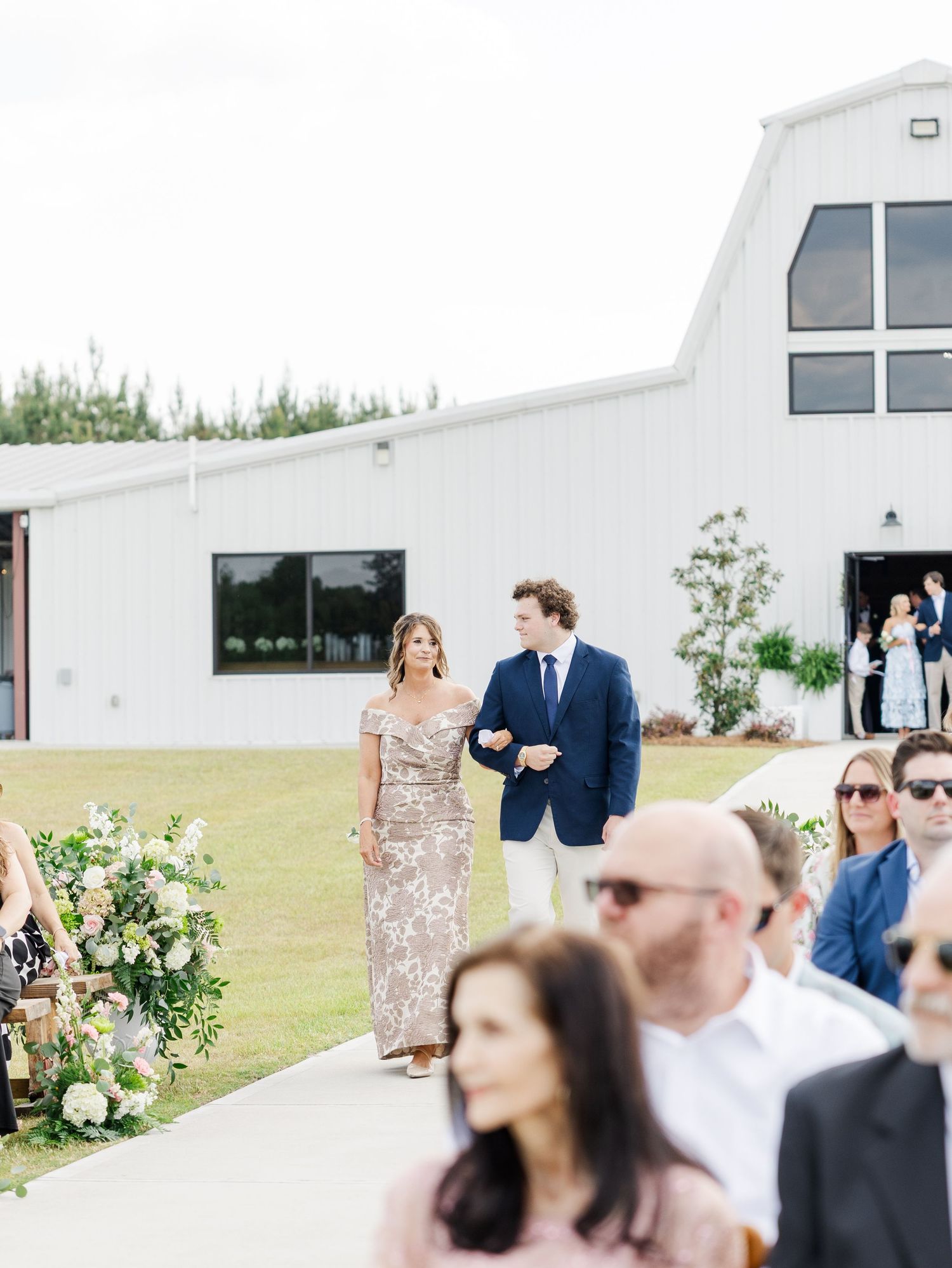 Mother of the groom in gold sequin gown being escorted down the aisle at an outdoor barn wedding ceremony.