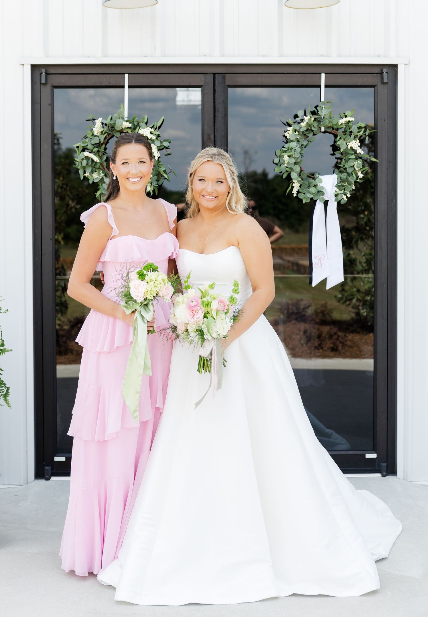 Bride in white gown and bridesmaid in pink dress holding bouquets in front of a white building with greenery wreaths.