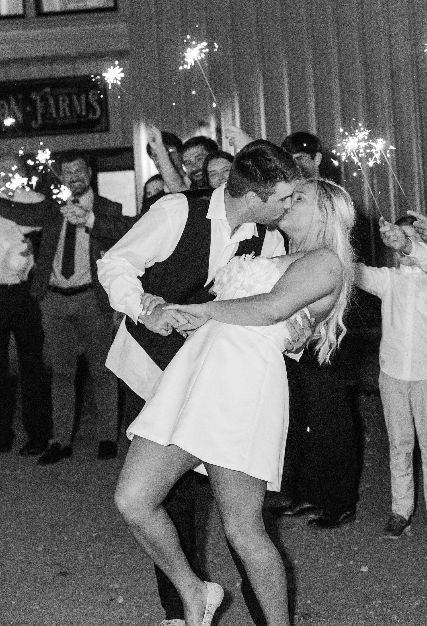 Newlyweds sharing a romantic kiss during a sparkler send-off surrounded by cheering wedding guests at night.