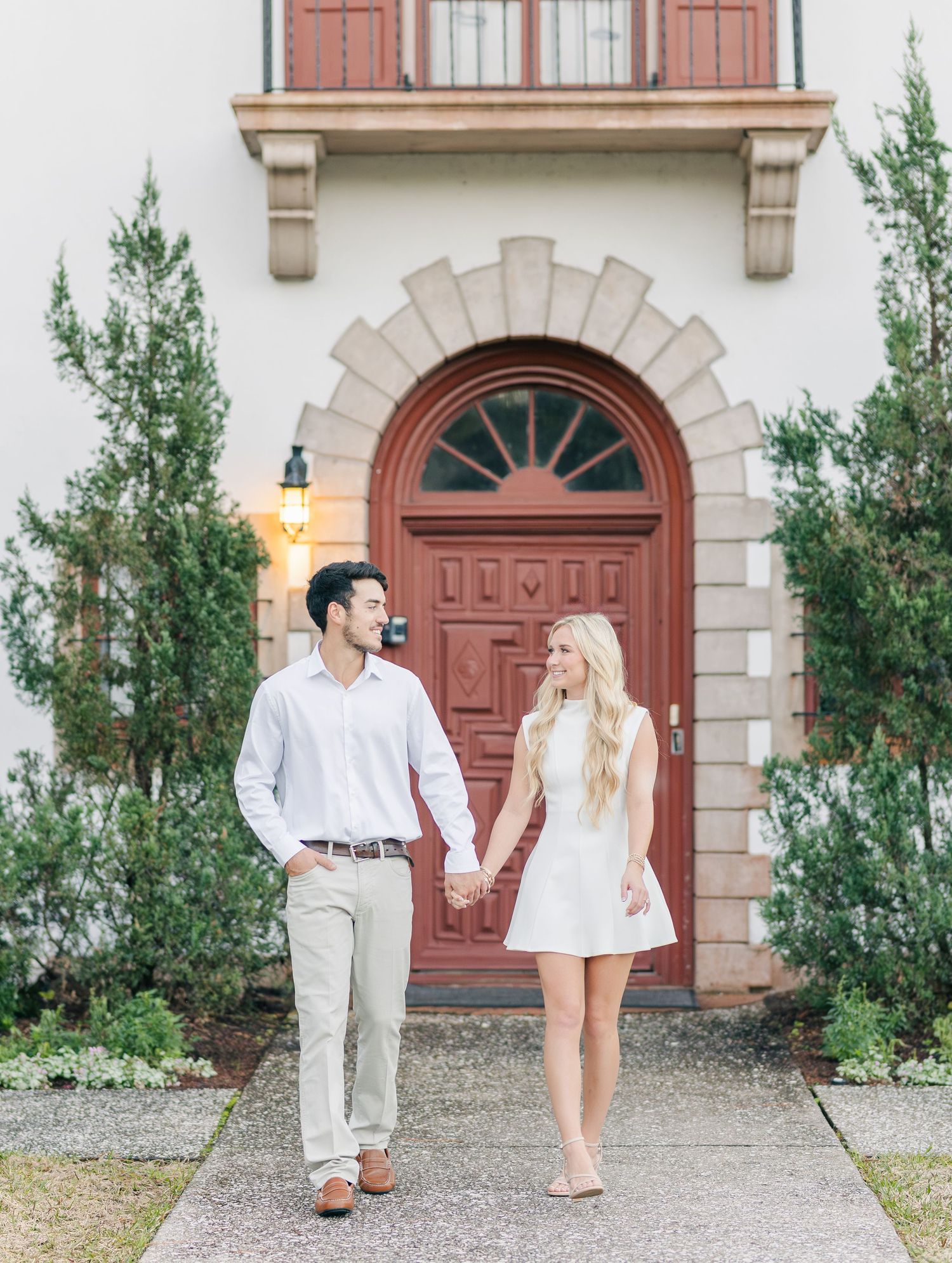 Smiling couple holding hands walking toward camera in front of elegant red arched door.