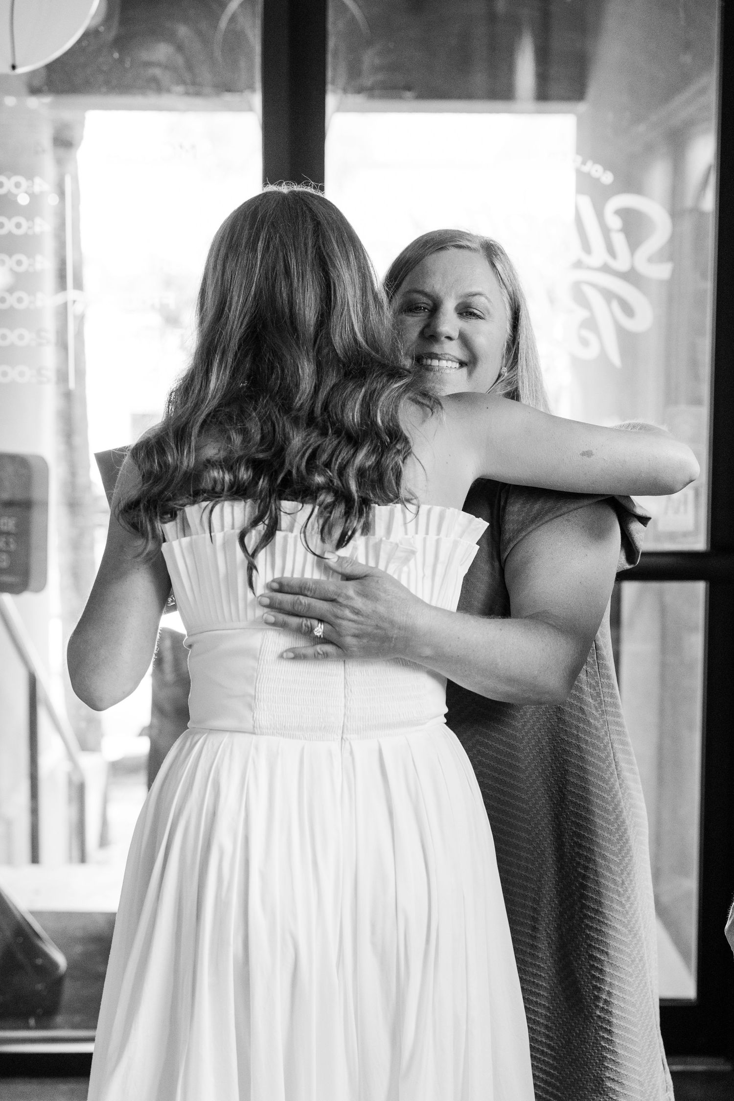 Black and white photo of a bride in a white gown sharing a warm hug with a smiling bridesmaid.