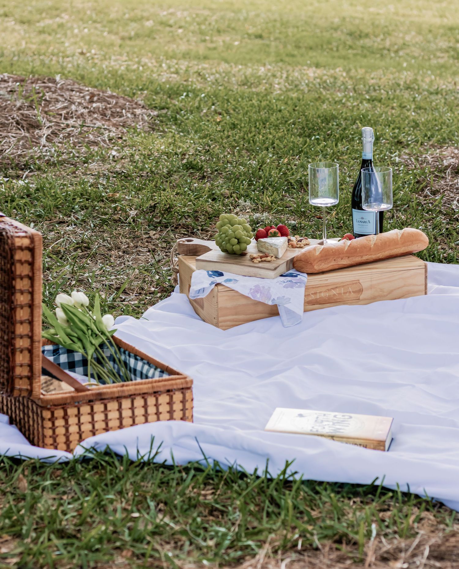Rustic outdoor picnic scene with wicker basket, wine, bread and wooden serving boards.