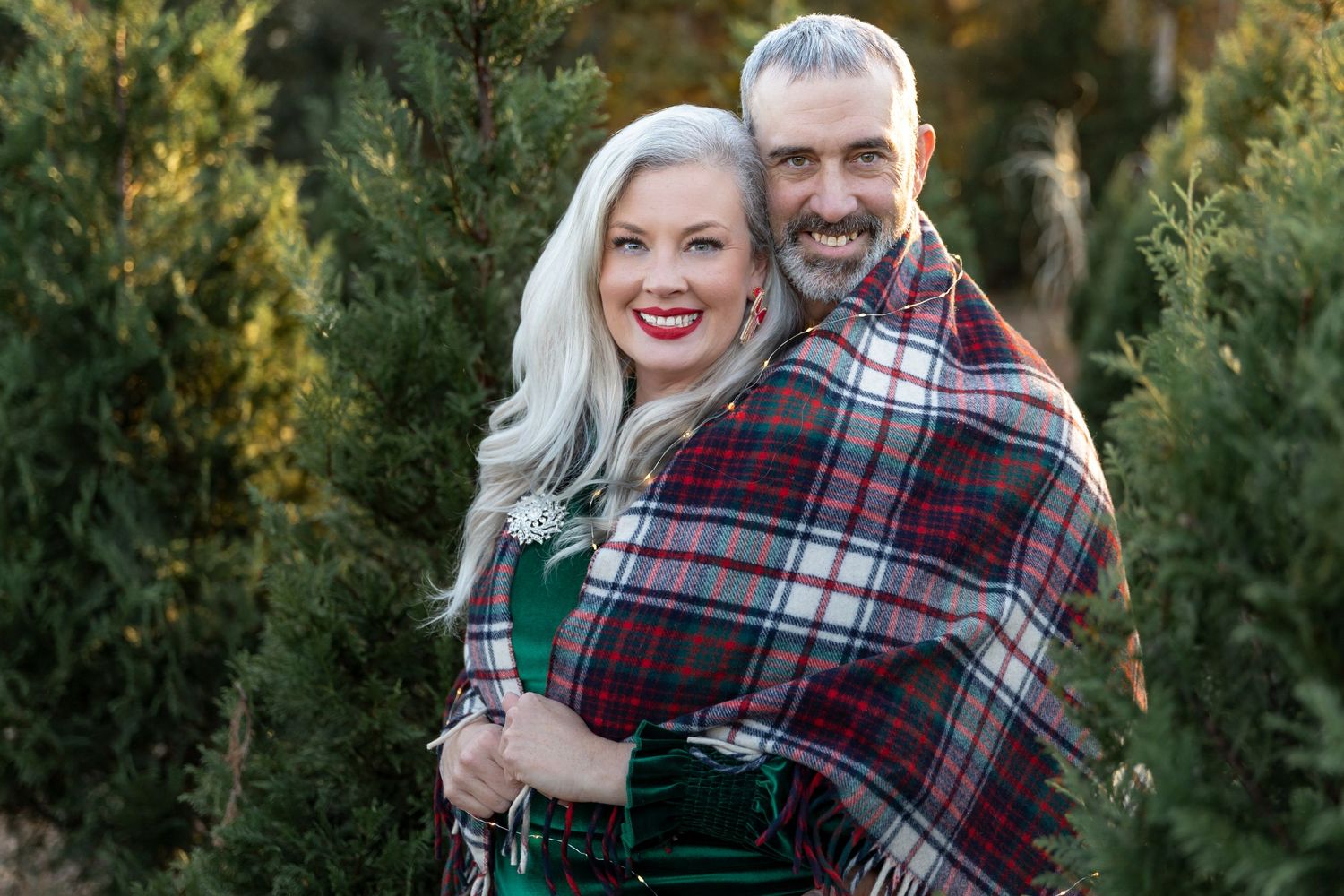 Couple wrapped in plaid blanket pose together in Christmas tree farm setting.