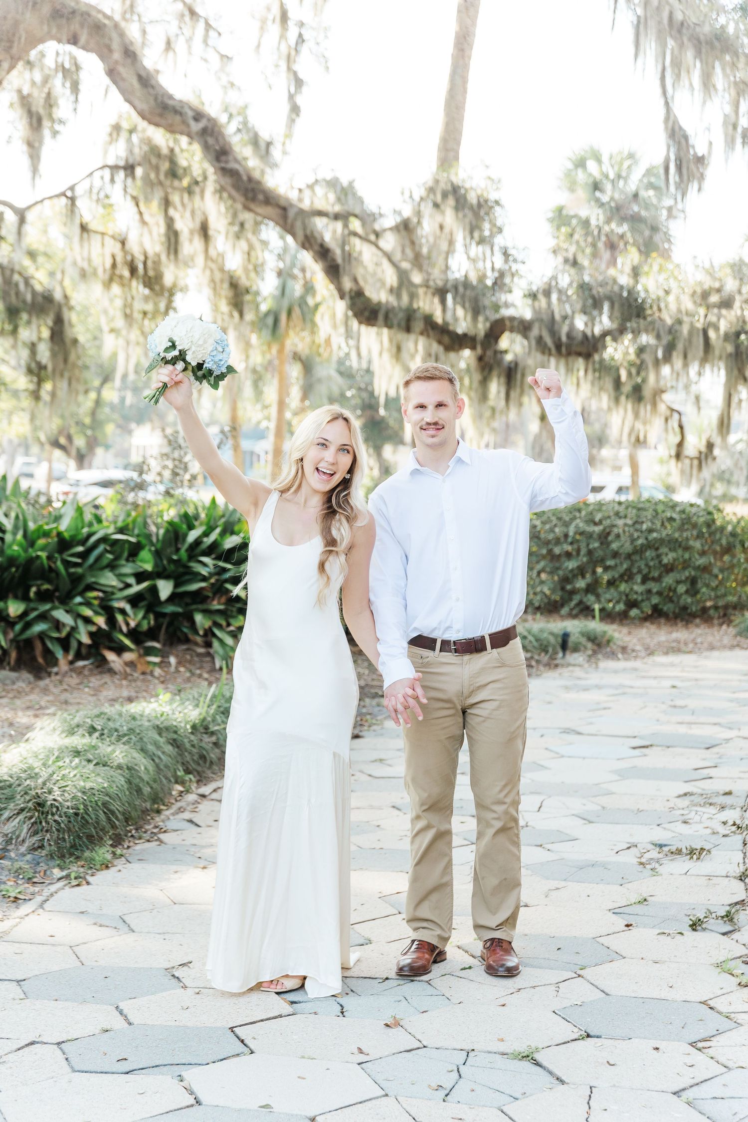 A joyful couple celebrates under Spanish moss trees while holding a bouquet.