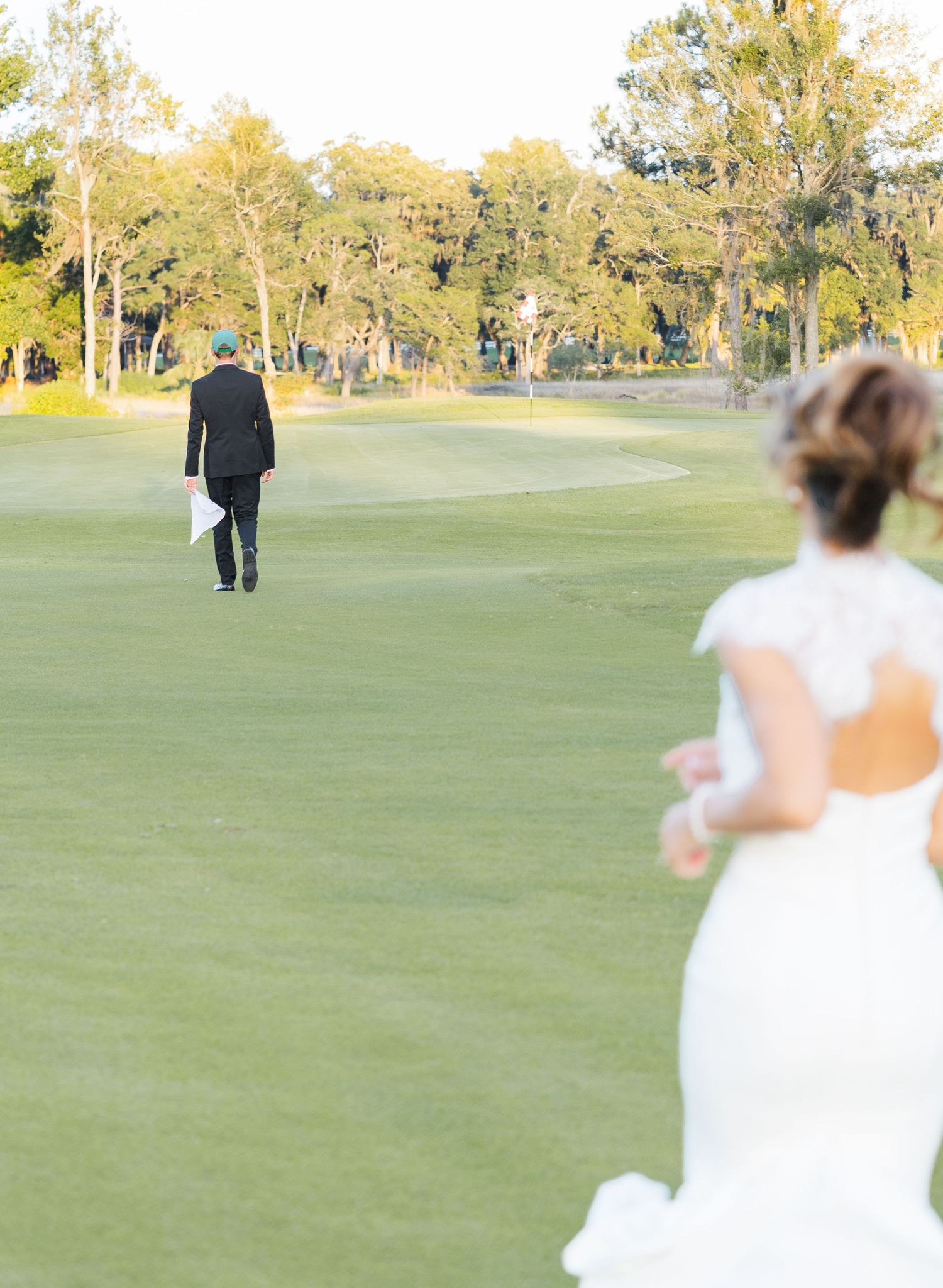 Bride in white gown running toward groom in black suit on lush green golf course at golden hour.