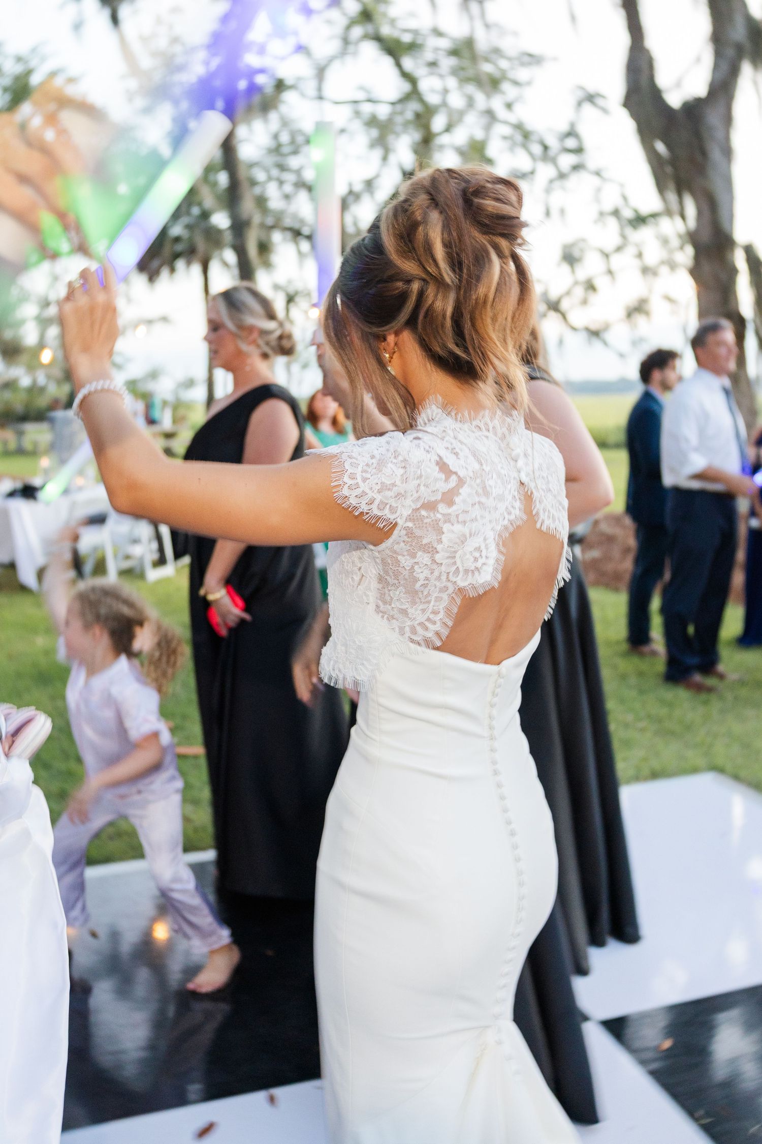 Back view of bride in lace open-back wedding gown dancing on outdoor dance floor with guests.