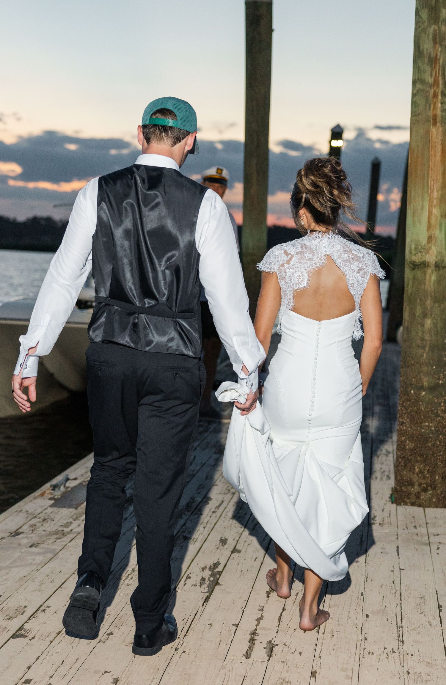 Bride and groom walk hand-in-hand on a dock at sunset, bride in lace-back gown, groom in vest and cap.