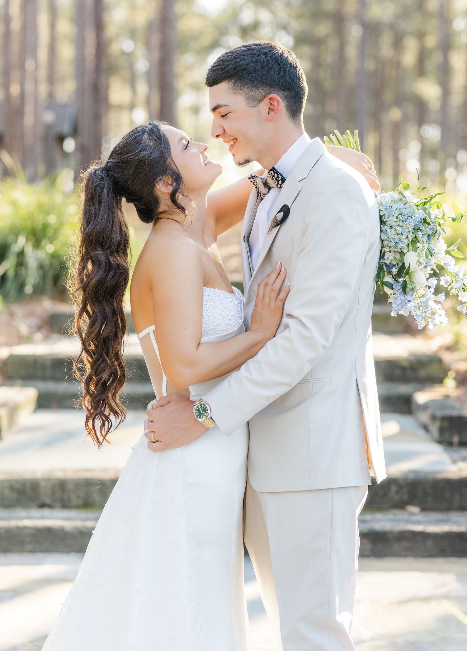 A bride and groom share a romantic moment outdoors, with the groom in a tan suit holding a white bouquet.