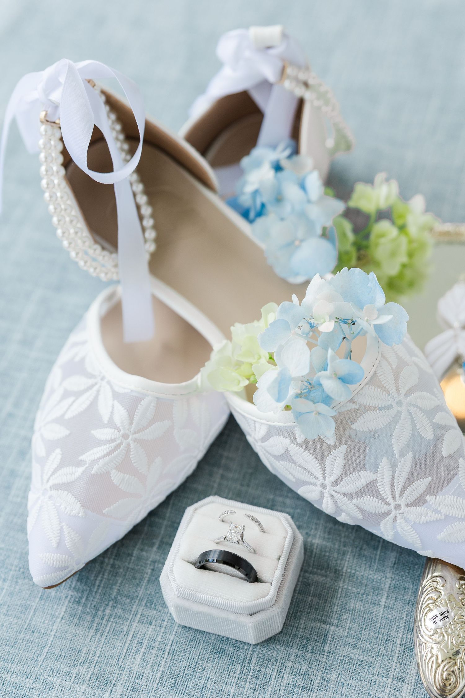 White lace bridal heels with pearl ankle straps beside blue hydrangeas and a wedding ring box on a blue surface.