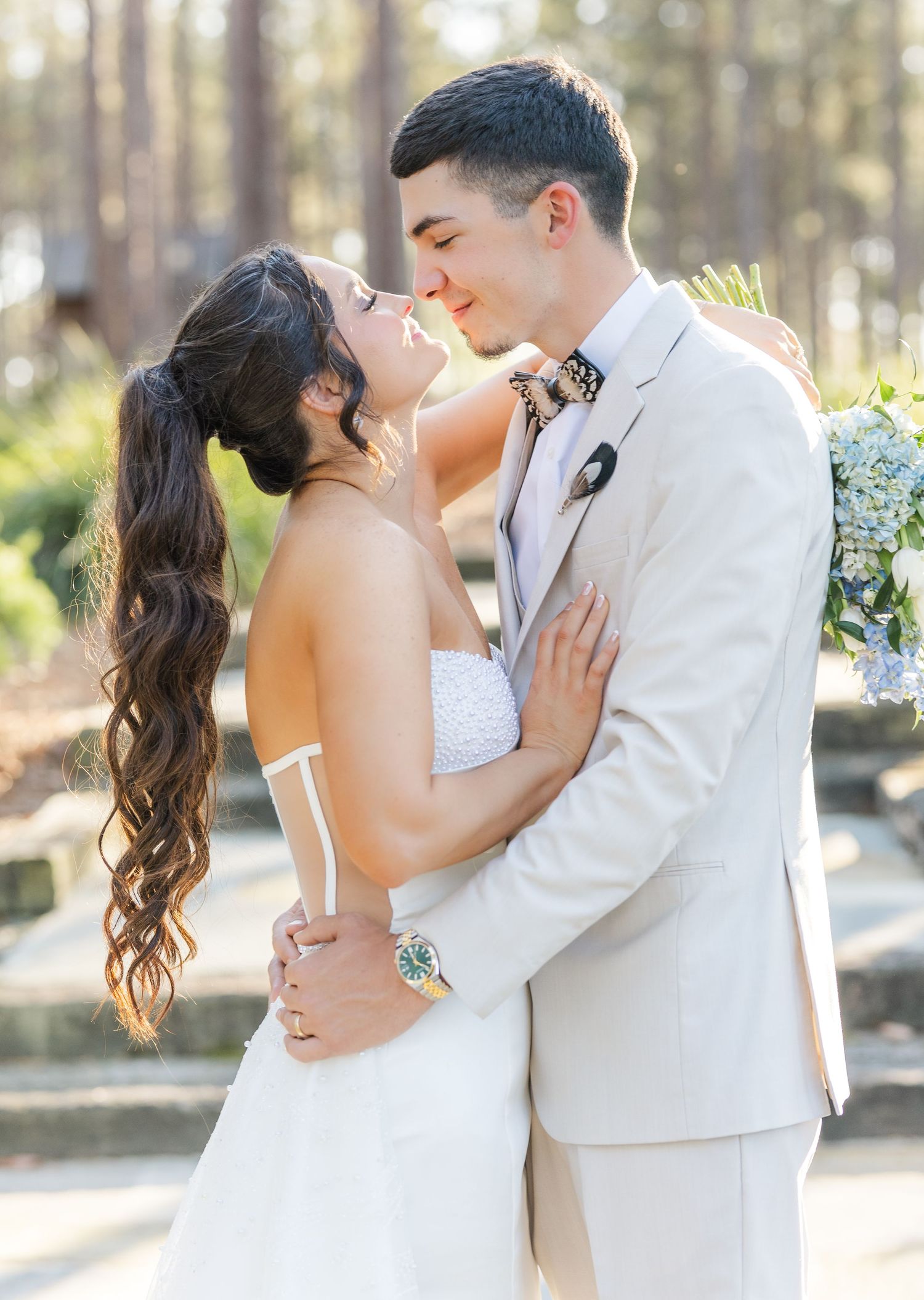 Bride and groom share a romantic almost-kiss moment outdoors, bride in strapless gown and groom in tan suit with bow tie.