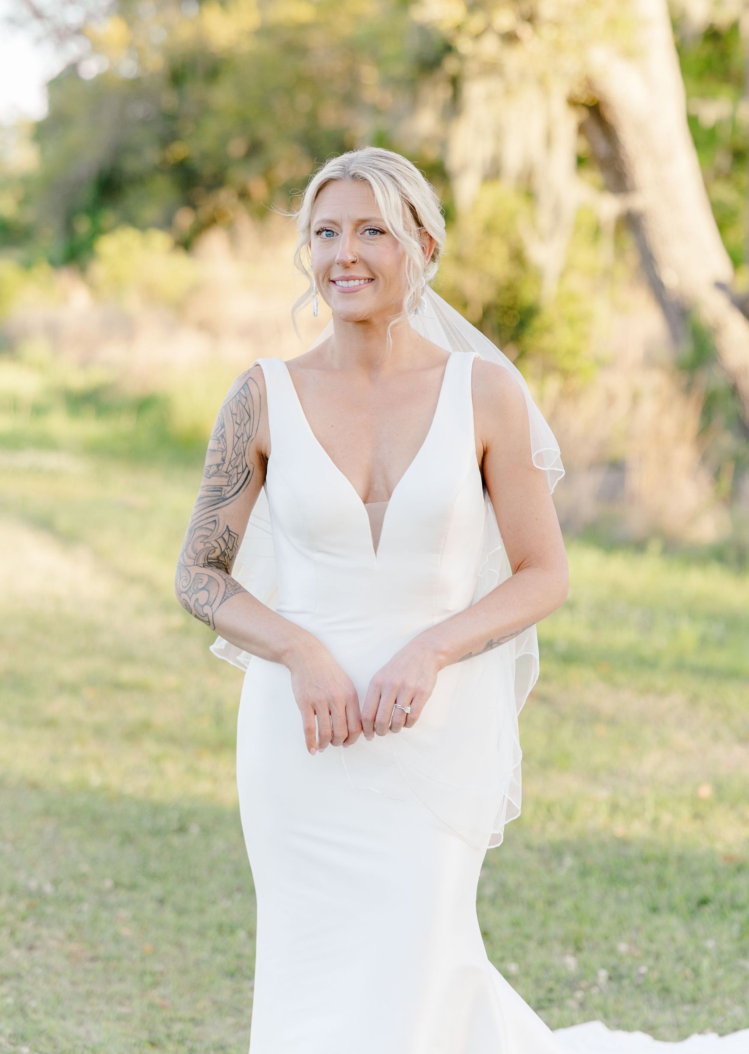 Smiling bride in elegant white V-neck wedding gown with tattoo sleeve and veil outdoors.