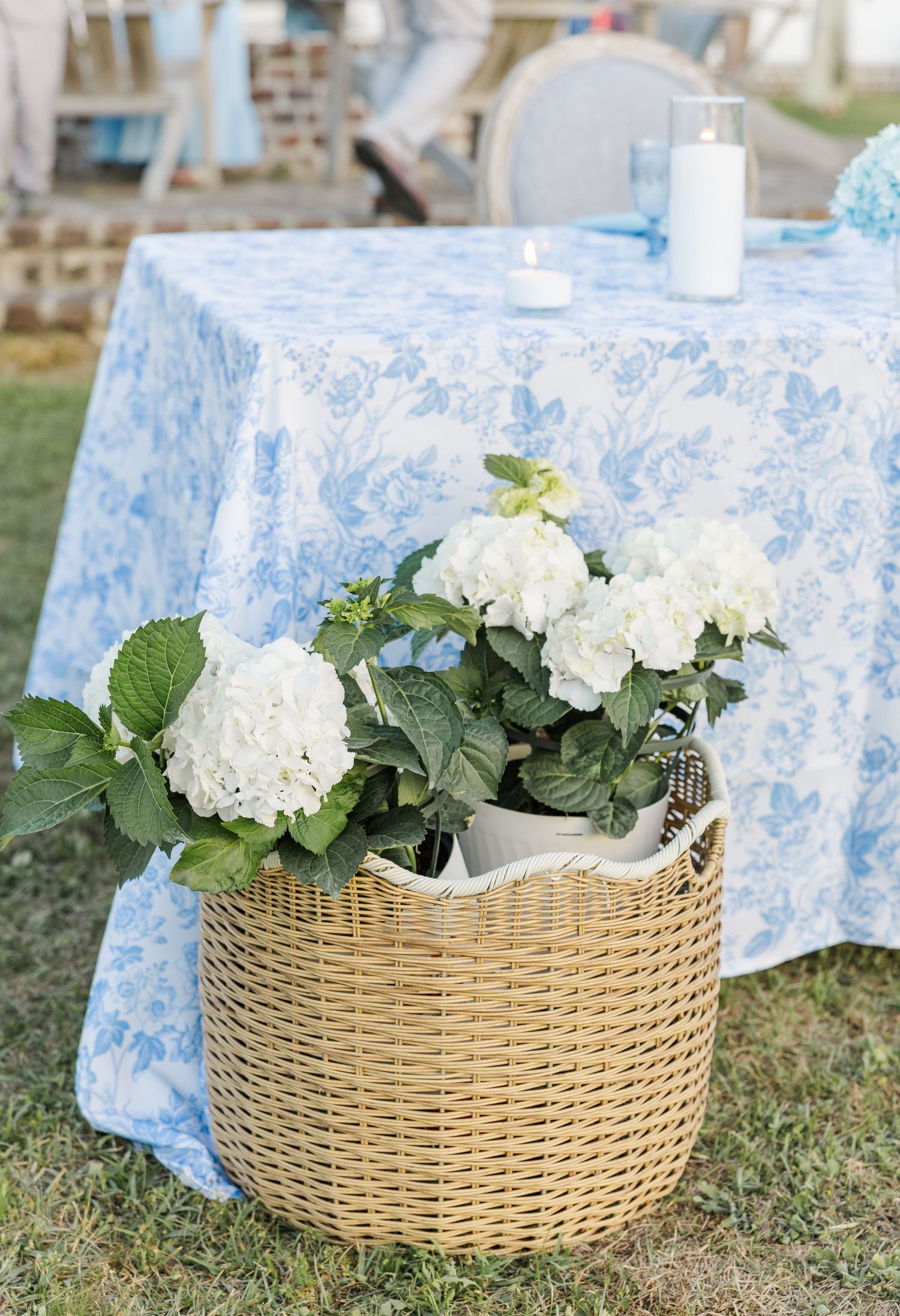 White hydrangeas in a wicker basket beside a blue floral tablecloth at an outdoor wedding reception.