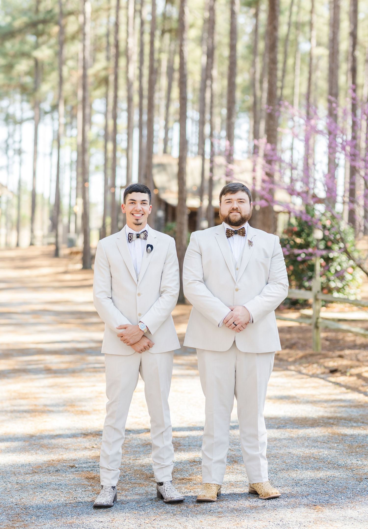 Two groomsmen in cream suits and bow ties pose on a pine tree-lined path at an outdoor wedding.