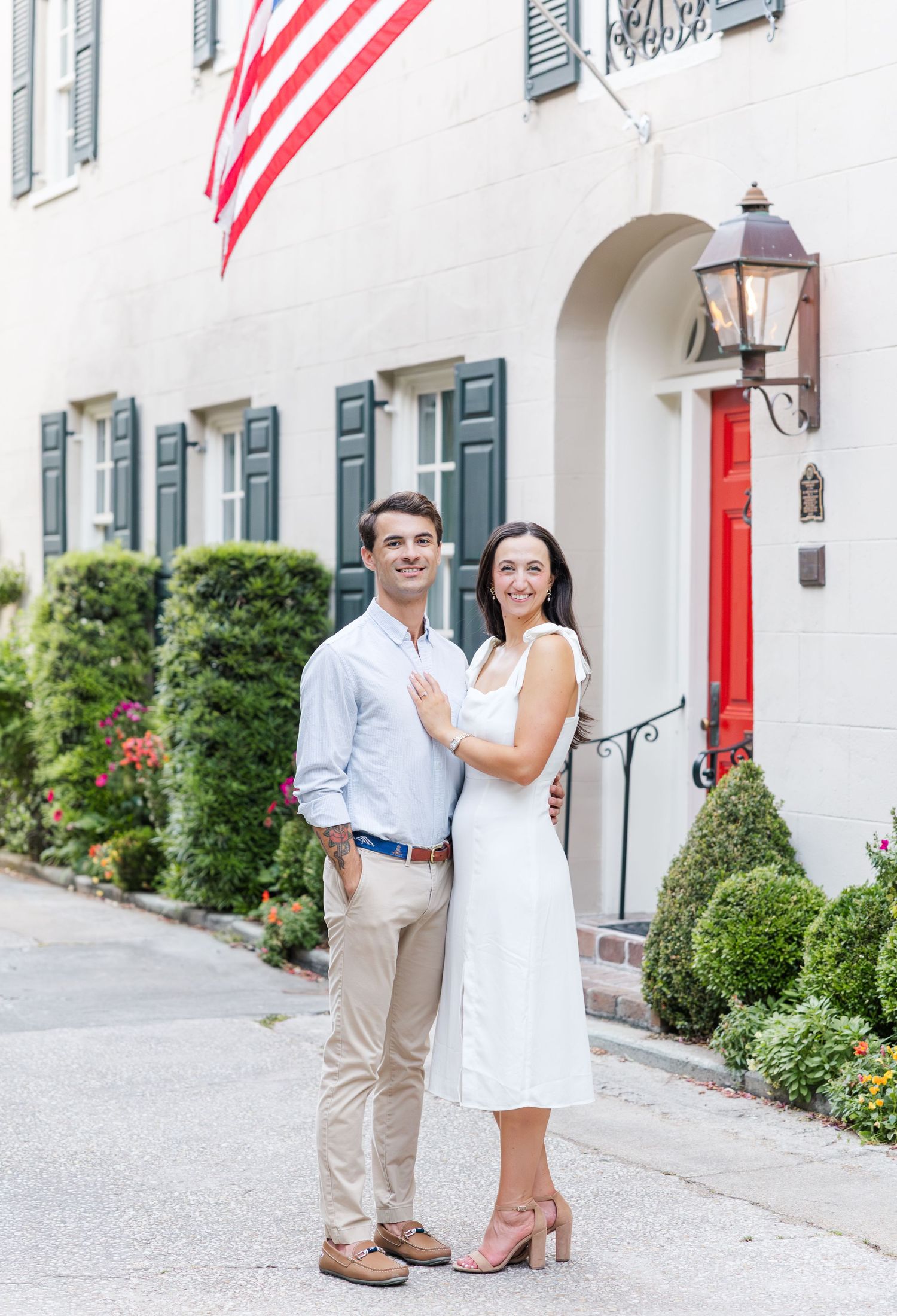 Couple posing in front of a white historic home with American flag and red door outdoors.