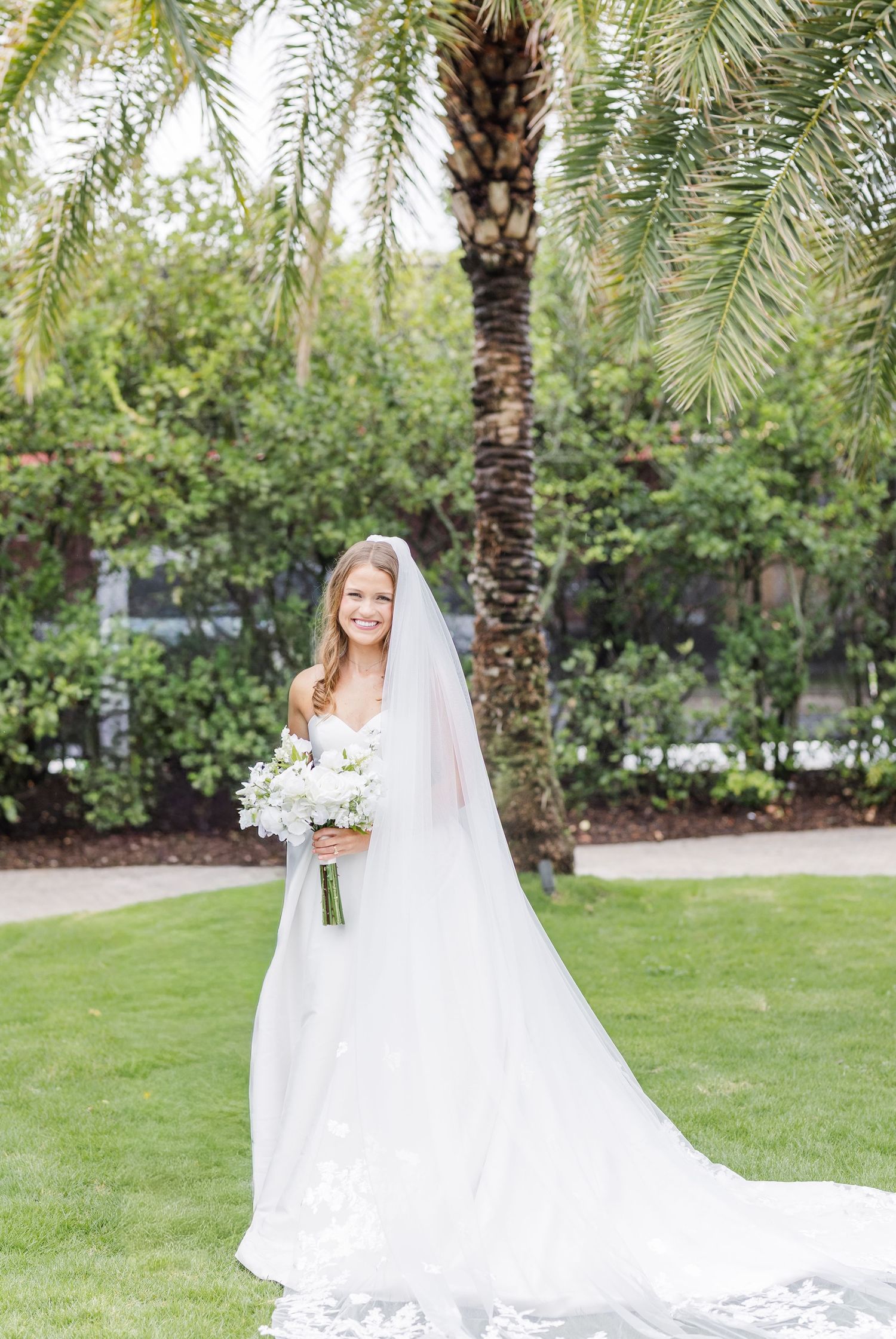 Smiling bride in white gown and veil holding white bouquet standing on green lawn near palm tree.