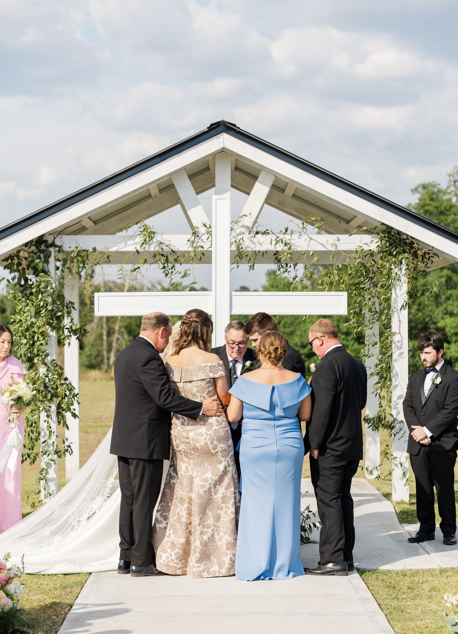Wedding ceremony under white pergola with cross as couple exchanges vows with family beside them.