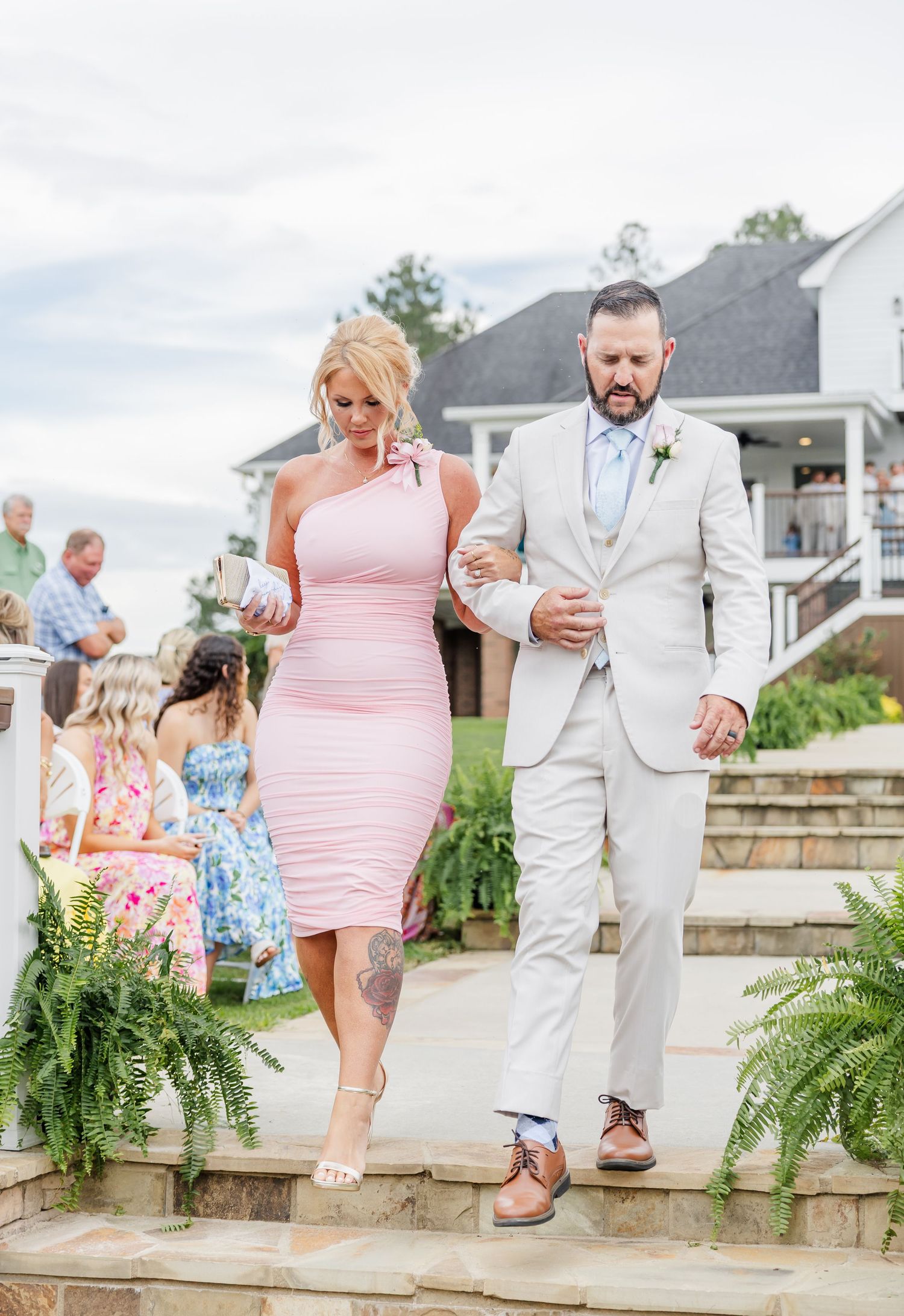 Woman in pink one-shoulder dress and man in tan suit descend stone steps at outdoor wedding venue.