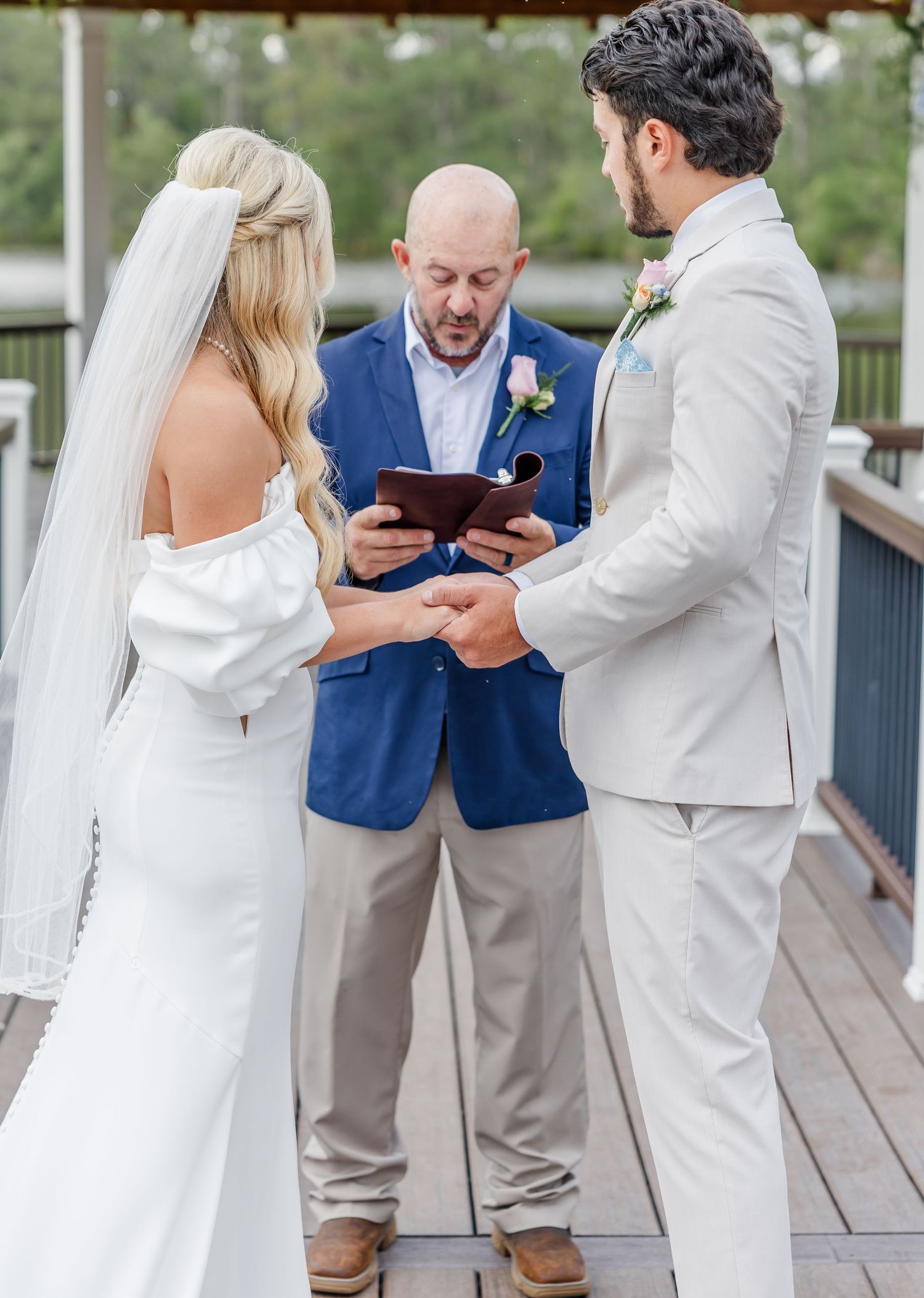 Officiant in navy blazer reads vows as bride and groom hold hands at outdoor wedding ceremony.