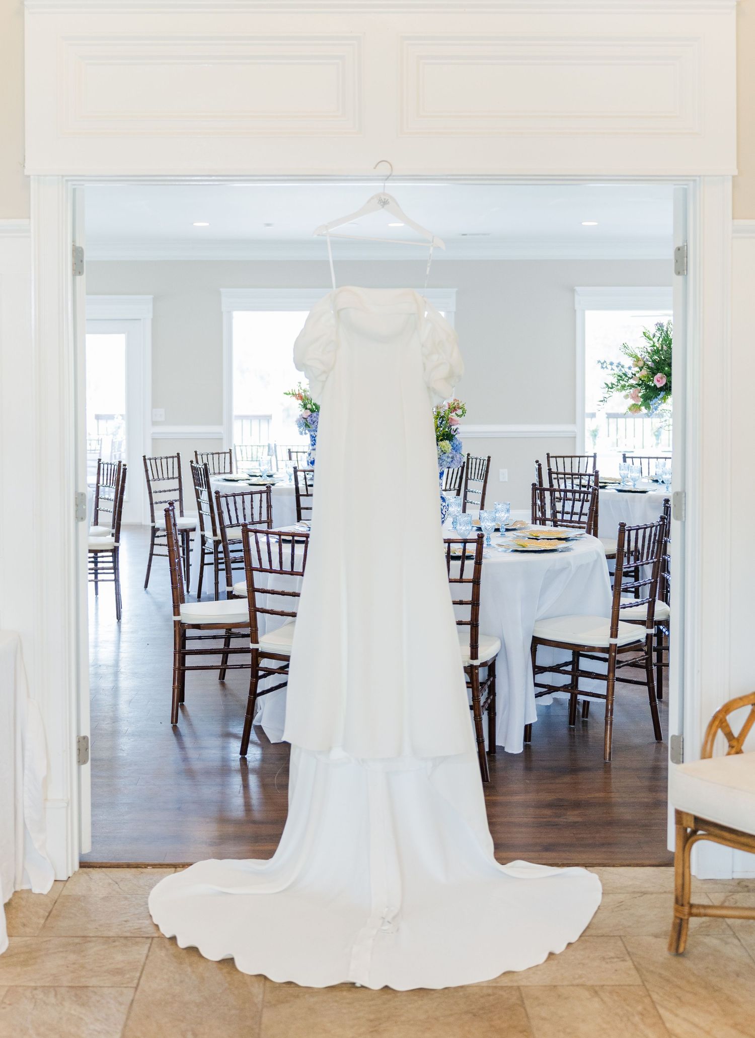 White wedding gown hanging in doorway of elegant reception venue with dark wood Chiavari chairs and white tablecloths.