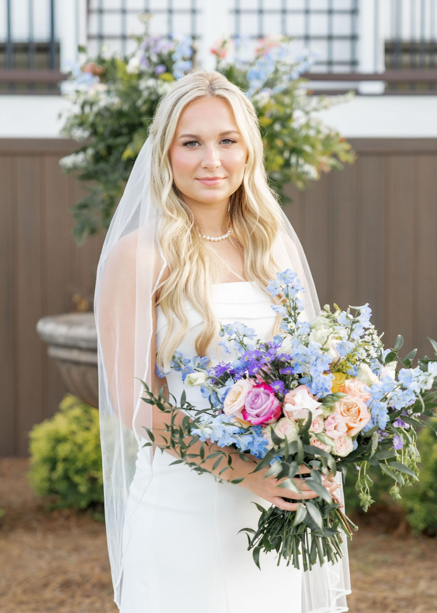 Blonde bride wearing cathedral veil holding blue, pink, and purple floral bouquet in front of venue exterior.