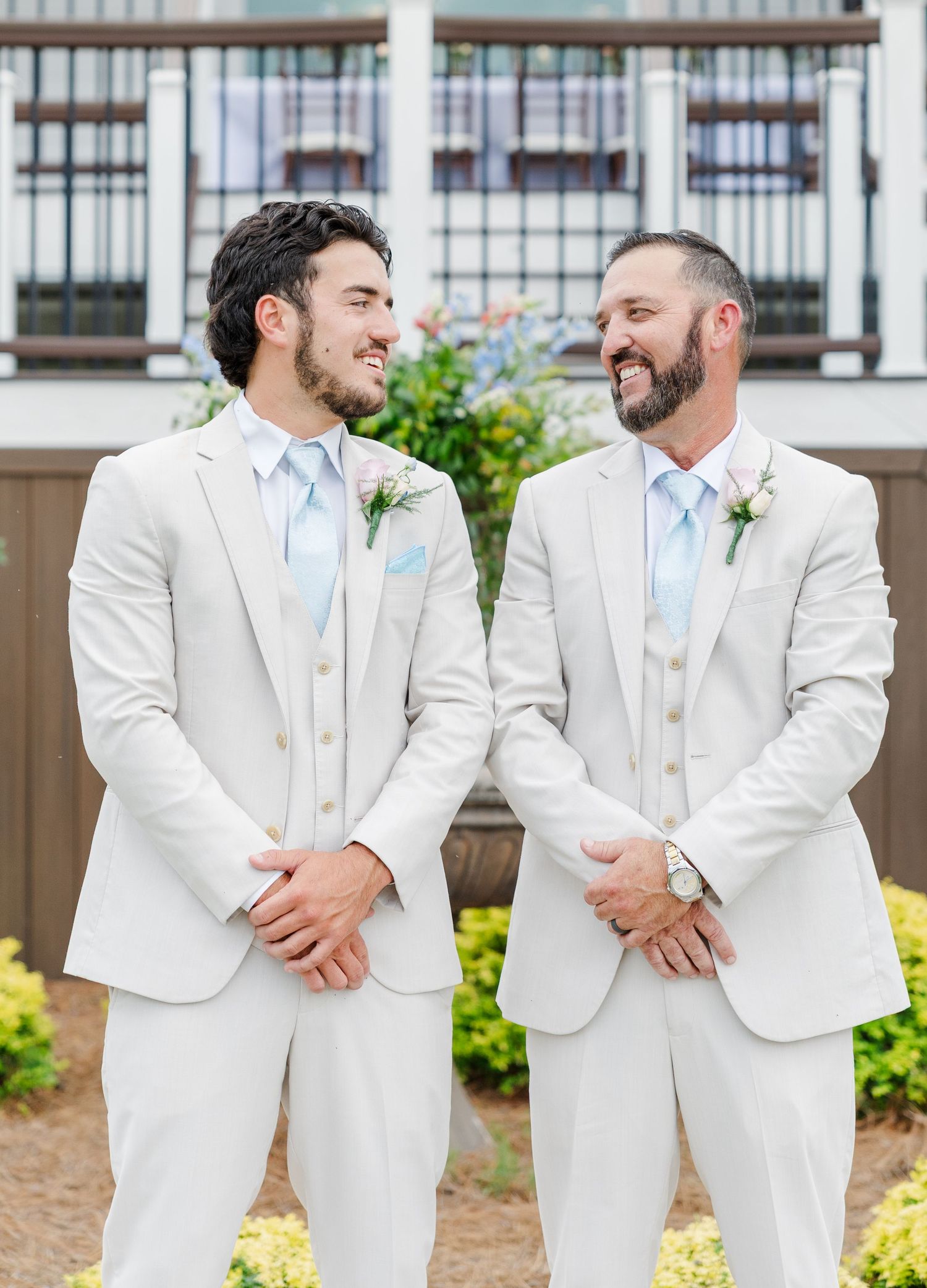 Two groomsmen in matching beige suits with blue ties and boutonnieres smiling at each other outdoors.