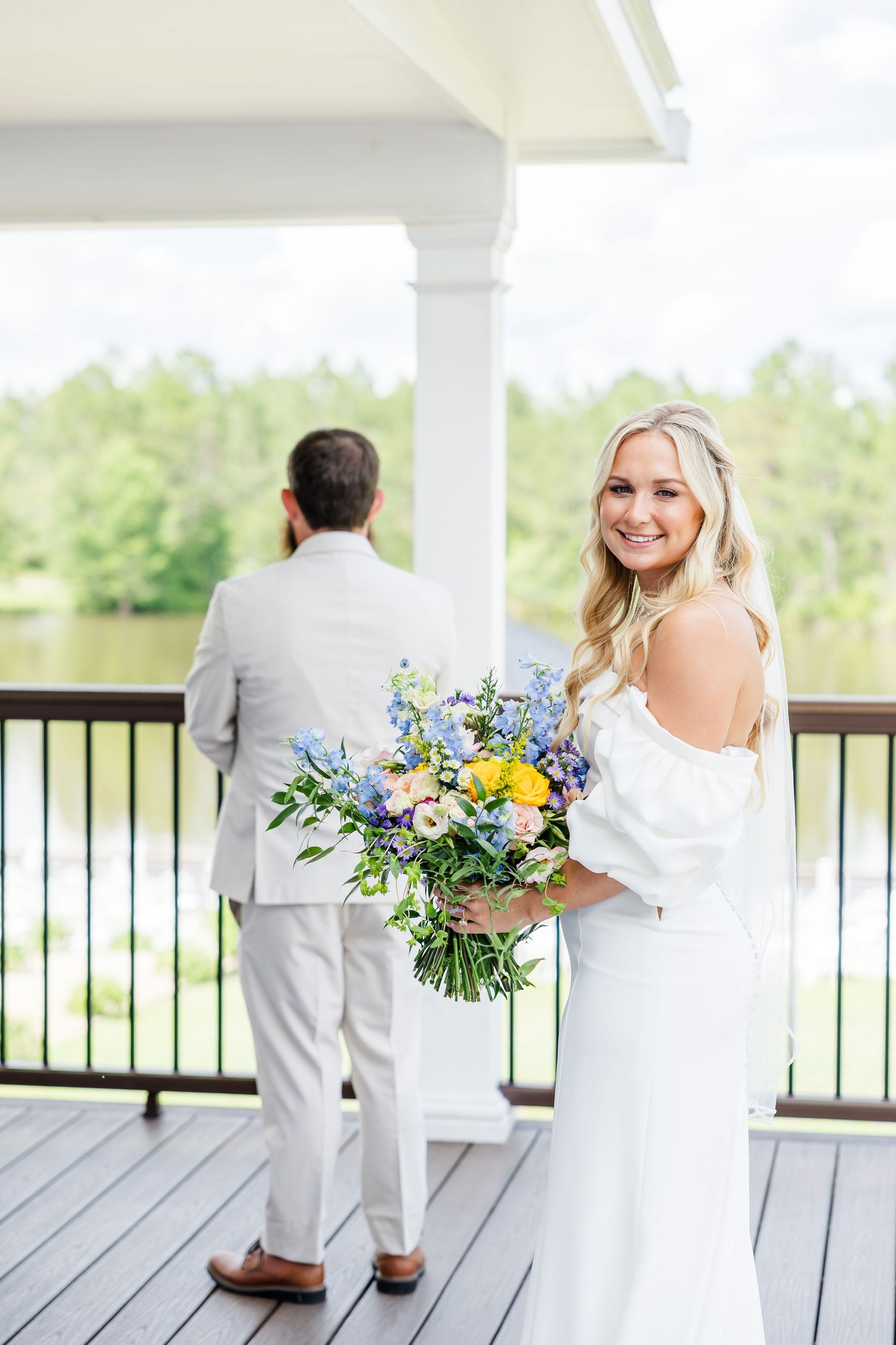 Smiling bride holding colorful bouquet during first look with groom on lakeside venue deck.
