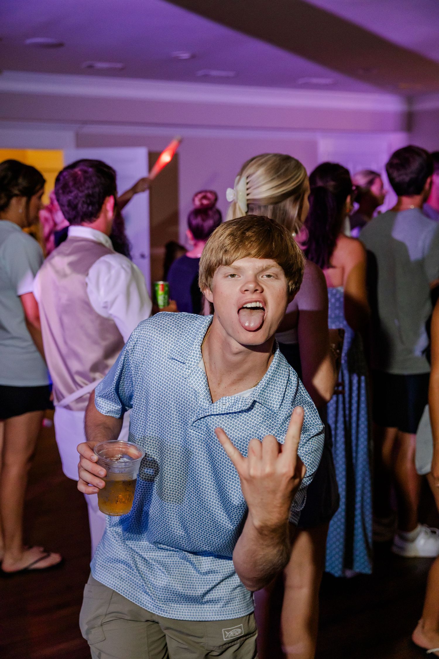 Young red-haired wedding guest making rock sign with tongue out while dancing at lively reception party.