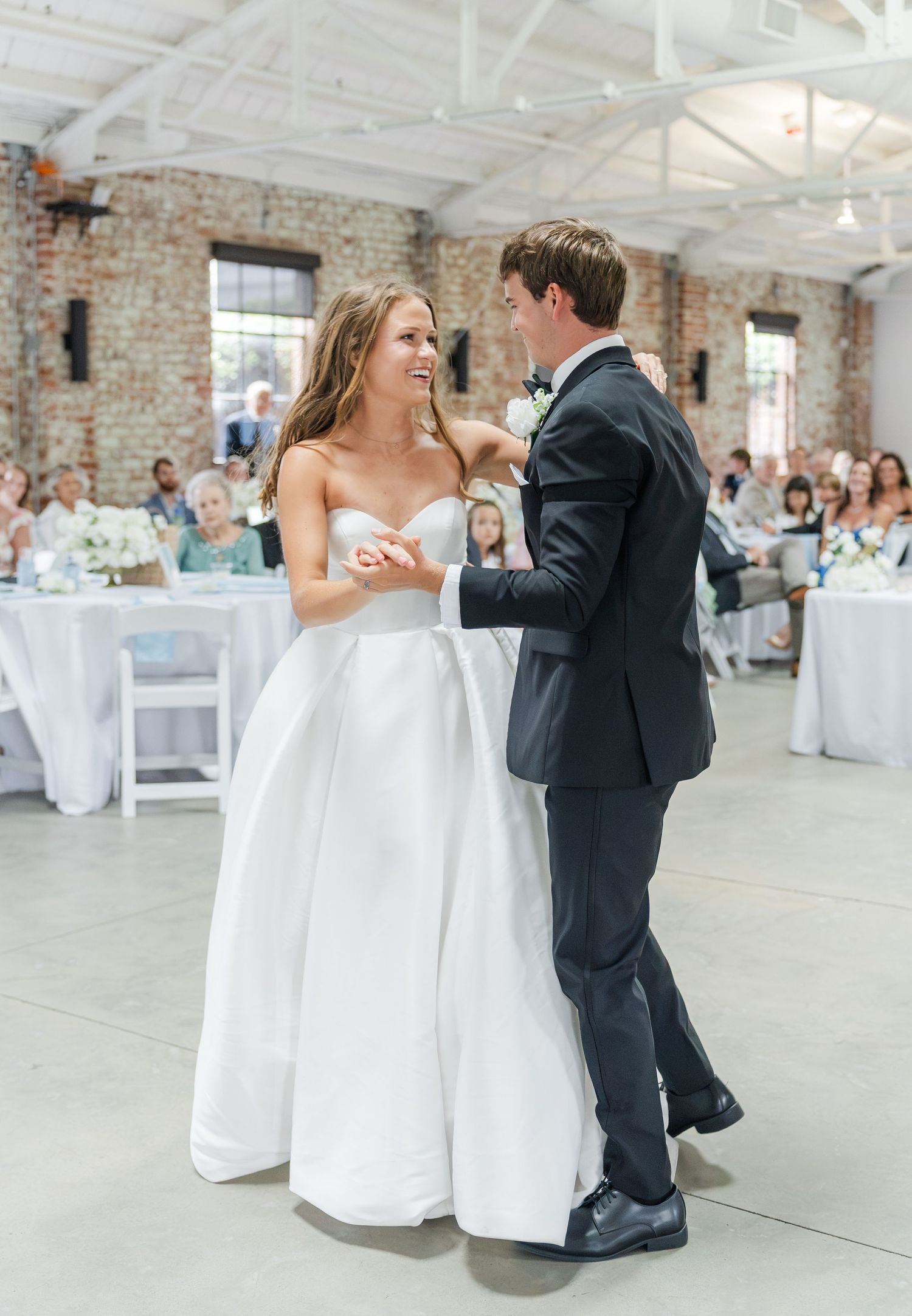 Smiling bride in strapless white gown dances with groom in black tuxedo at wedding reception.