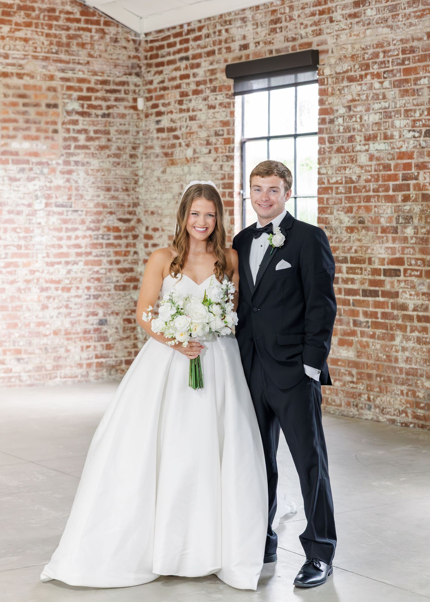 Smiling bride in white ballgown and groom in black tuxedo posing in rustic exposed brick venue.