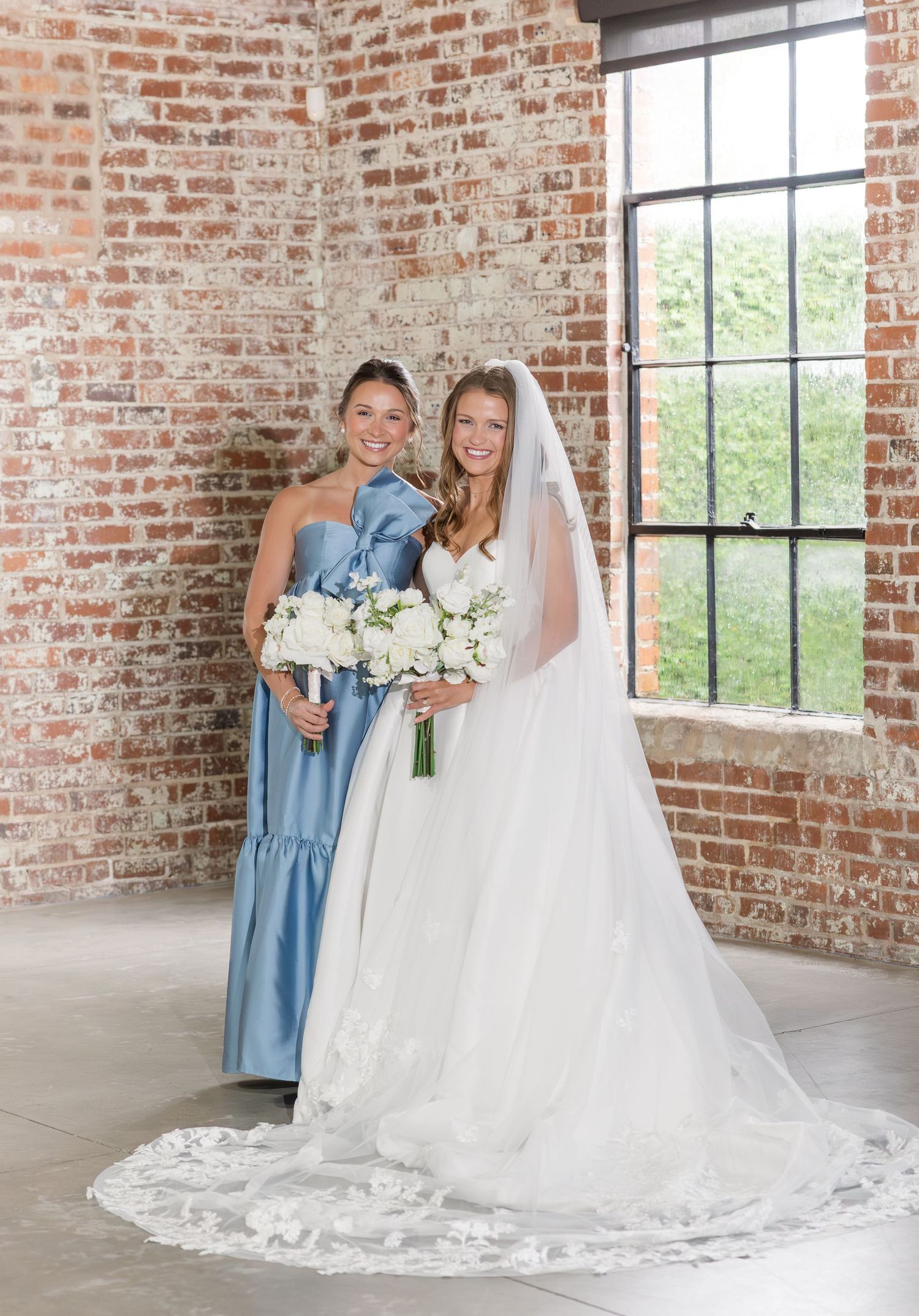 Bride in white ball gown with veil poses with bridesmaid in blue satin dress against rustic brick wall.