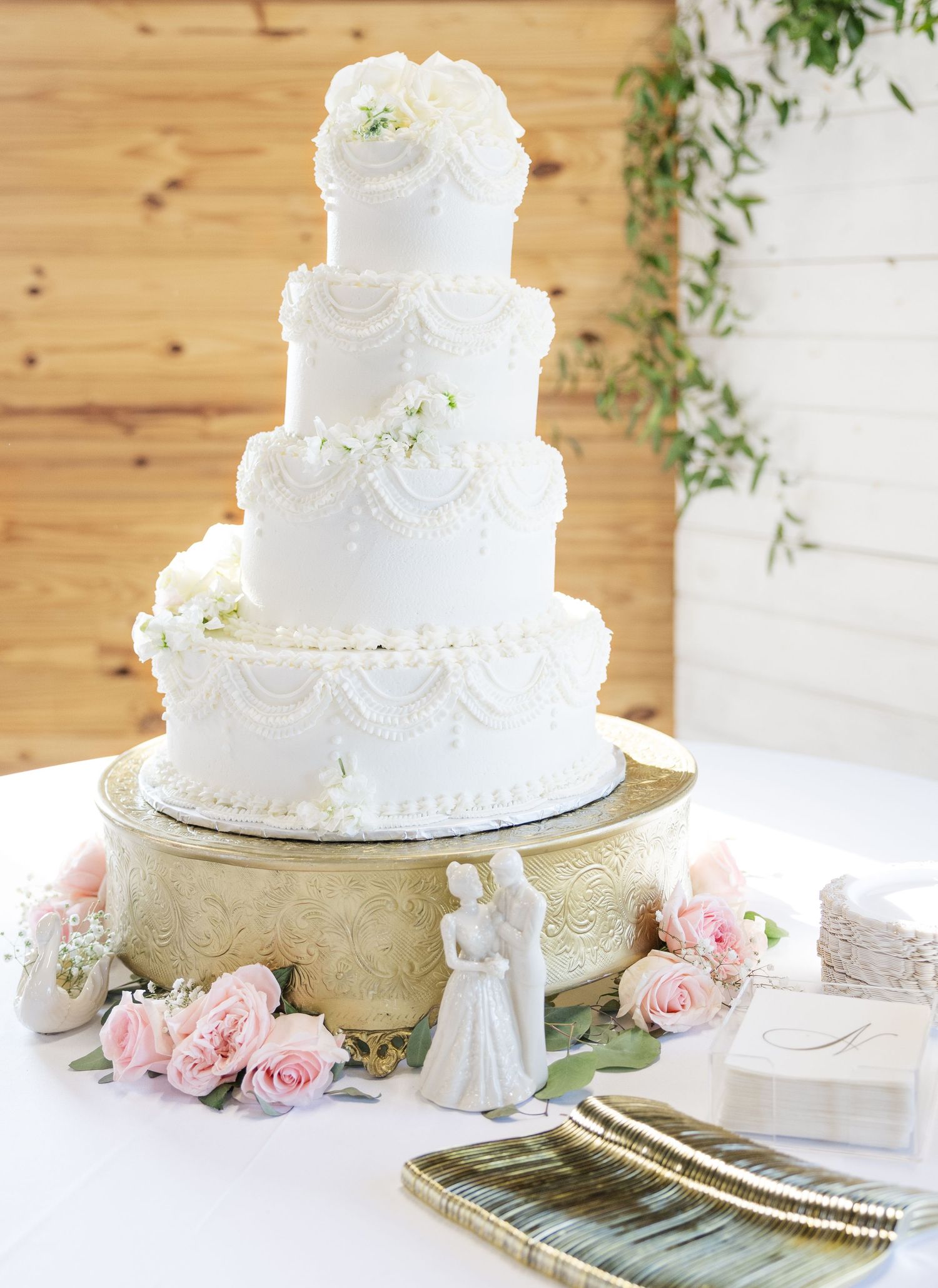 Four-tier white wedding cake with floral details on gold stand surrounded by pink roses.
